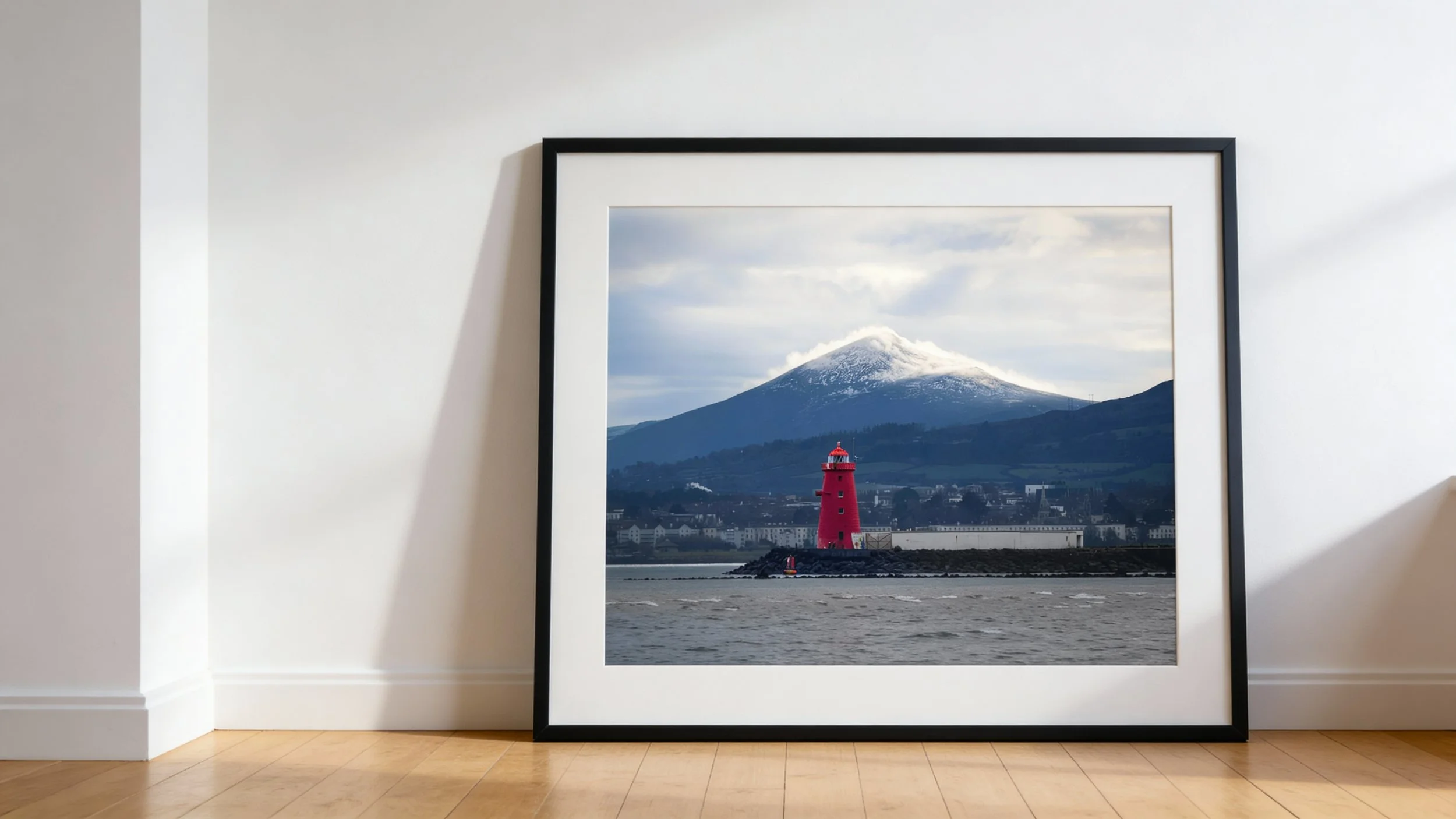 Framed photograph of a red lighthouse along a coastline with snow-capped mountains and cloudy sky in the background, leaning against a white wall in a room with wooden flooring.