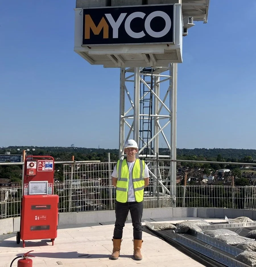 A man wearing a white hard hat, a high-visibility vest, and brown work boots, standing on a rooftop next to a red fire extinguisher and a red fire alarm box. Behind him is a tall metal structure with a large sign reading 'MYCO', and a cityscape under a clear blue sky.