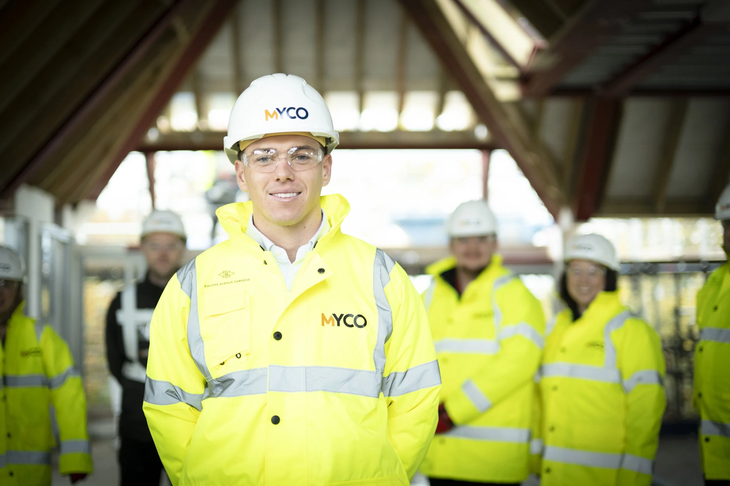 Construction workers wearing yellow vests and white helmets gathered around a worker giving instructions at a building site.
