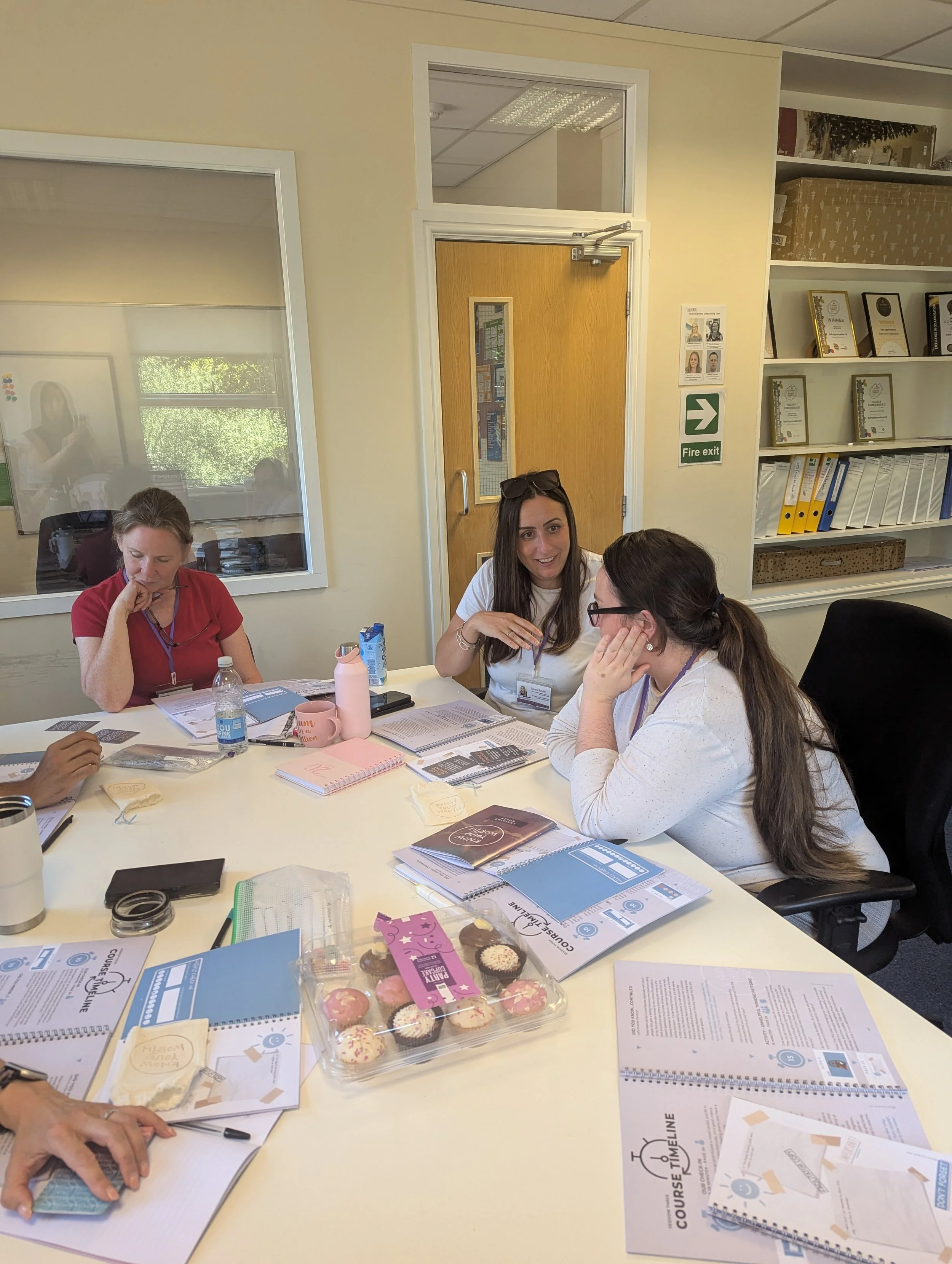 Women seated around a table in a meeting room, engaged in conversation, with notebooks, documents, snacks, and water bottles on the table, and a window and bookshelf in the background.
