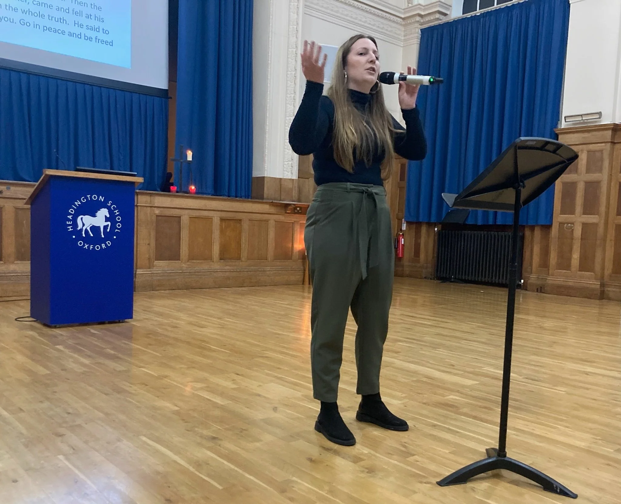 A woman standing on a wooden stage, speaking into a microphone, with a music stand in front of her. She is dressed in a black turtleneck, olive green pants, and black shoes. Behind her are blue curtains, a lectern with the Headington School Oxford lo
