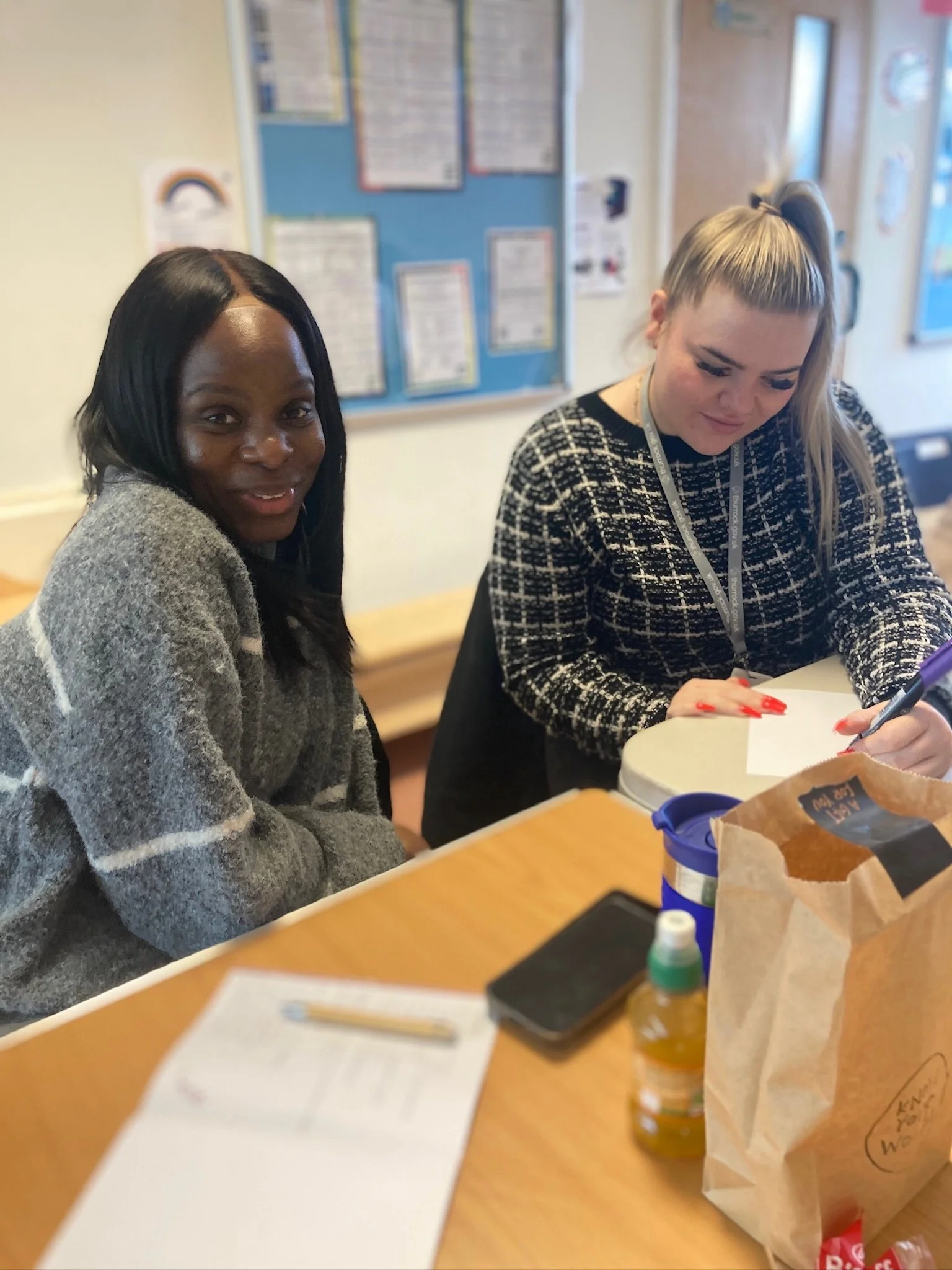 Two women sitting at a table in a classroom; one is writing on a notepad, the other is looking at the camera.