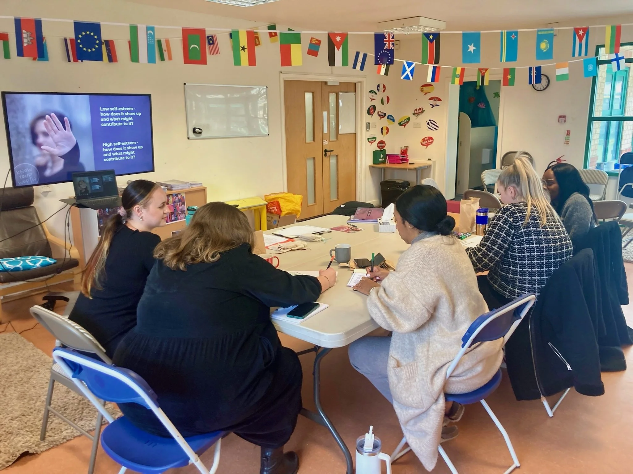 A group of six women seated around a conference table in a classroom or meeting room, engaged in a meeting or discussion. There is a large screen on the wall displaying a presentation, and the room is decorated with international flags and colorful w