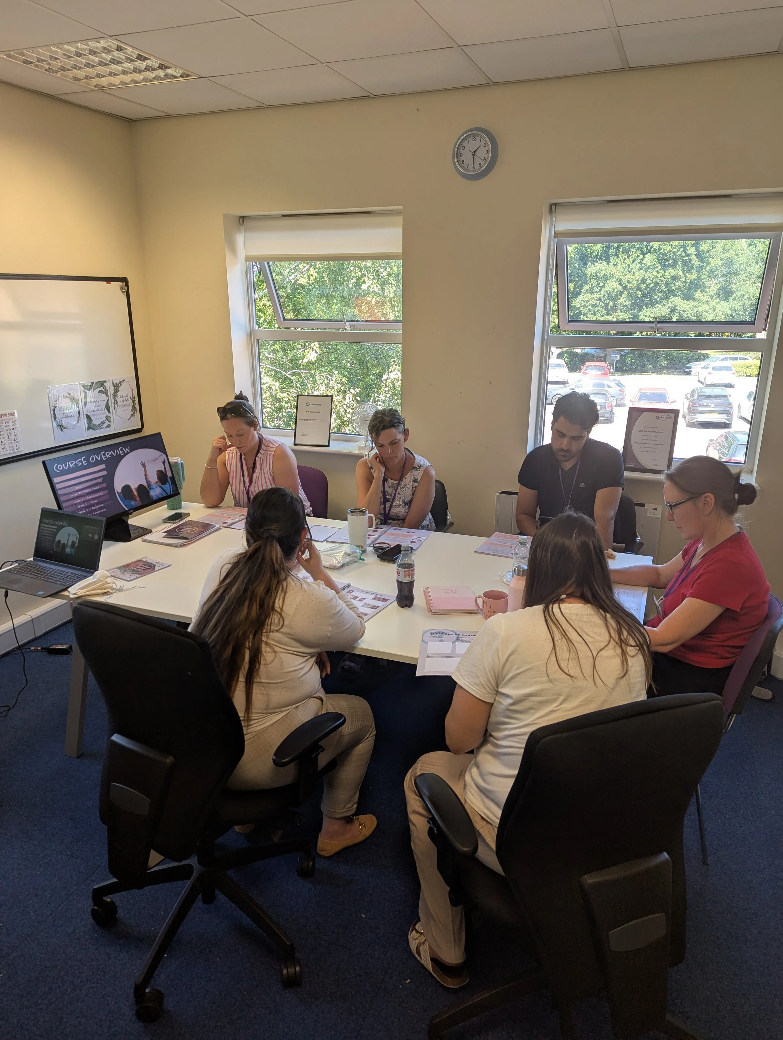 Group of six people sitting around a rectangular table in a classroom or meeting room, looking at papers and a monitor with a presentation. The room has two windows with open blinds, showing parked cars outside. A whiteboard and a clock are on the wa