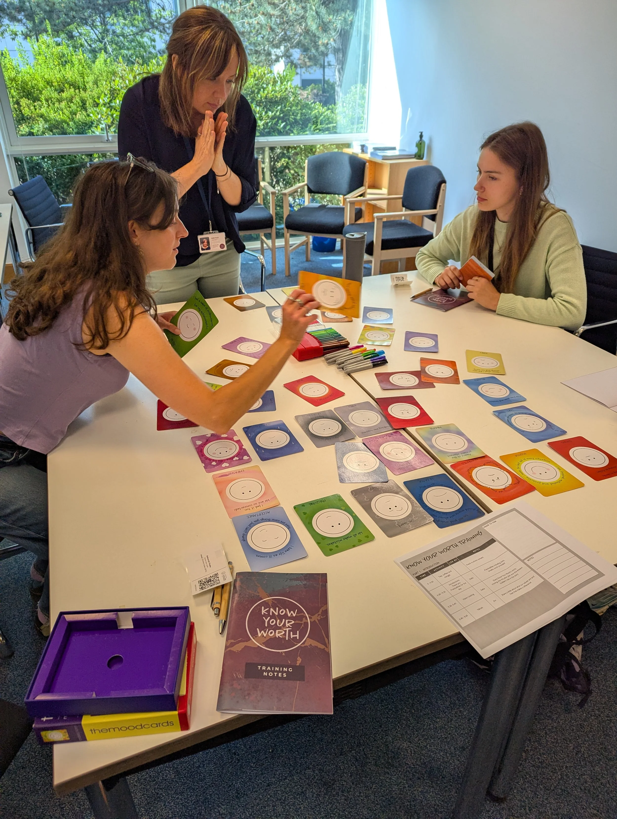 Three women playing a card game called 'Know Your Worth' in a conference room with large windows and a view of greenery outside.