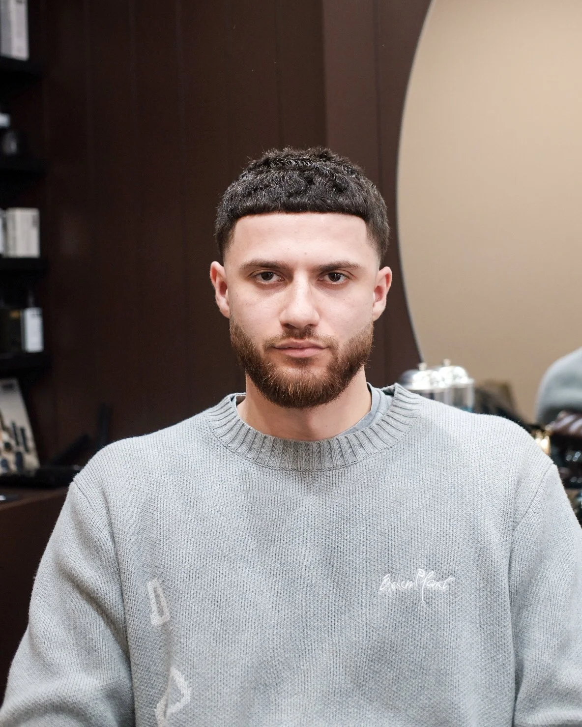 Man sitting in a barber shop with a fresh haircut and beard trim, wearing a light gray sweater.