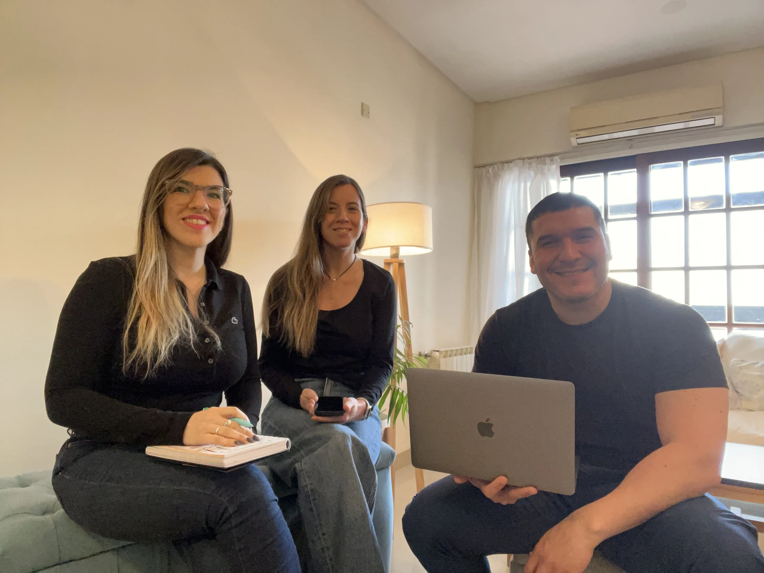 Tres personas sentadas en un sofá, dos mujeres y un hombre, en una sala con luz natural y una lámpara, sonriendo y usando dispositivos electrónicos.