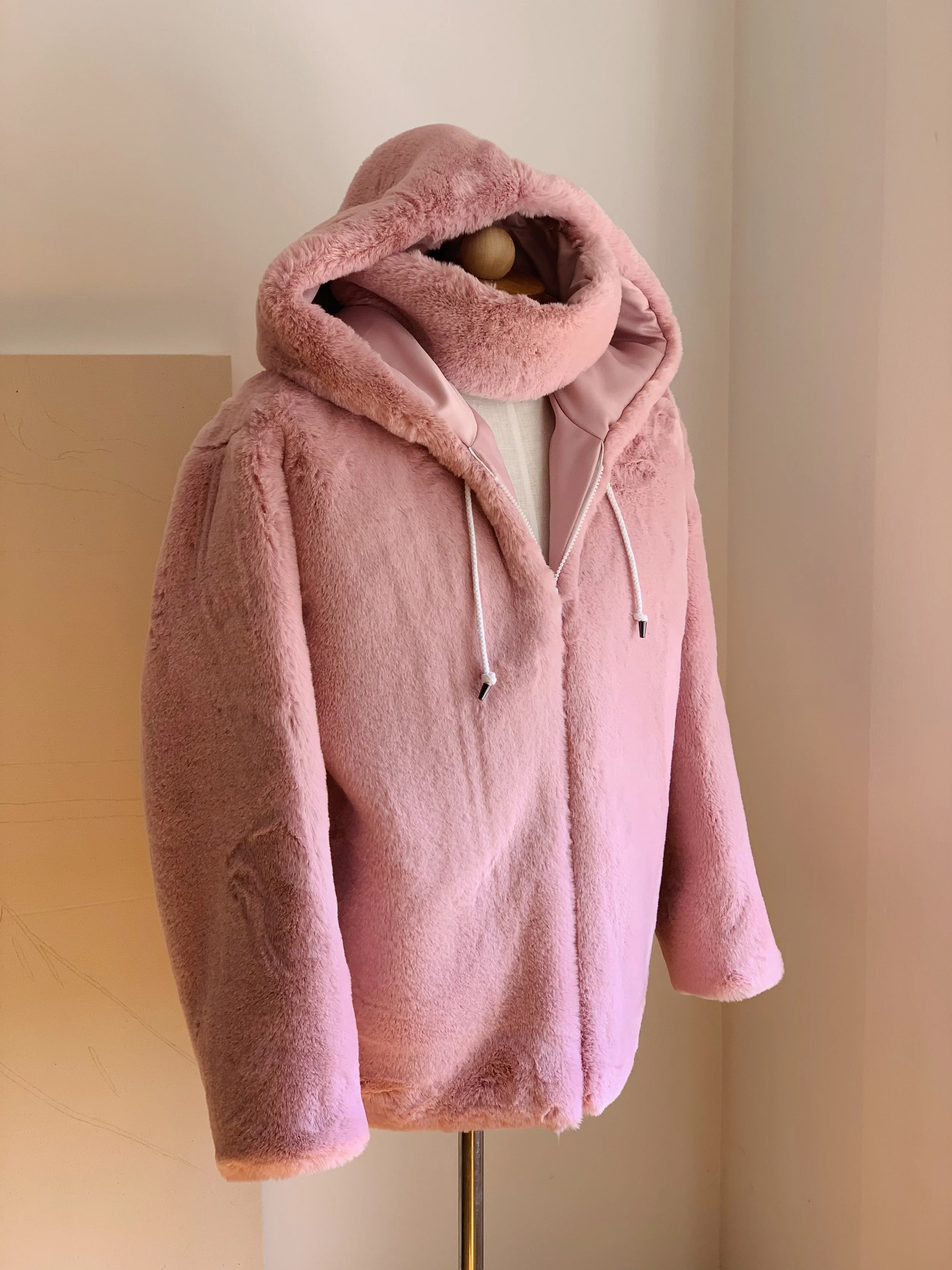 A pink faux fur hoodie with a large hood, open front, and white drawstrings displayed on a mannequin against a beige wall.