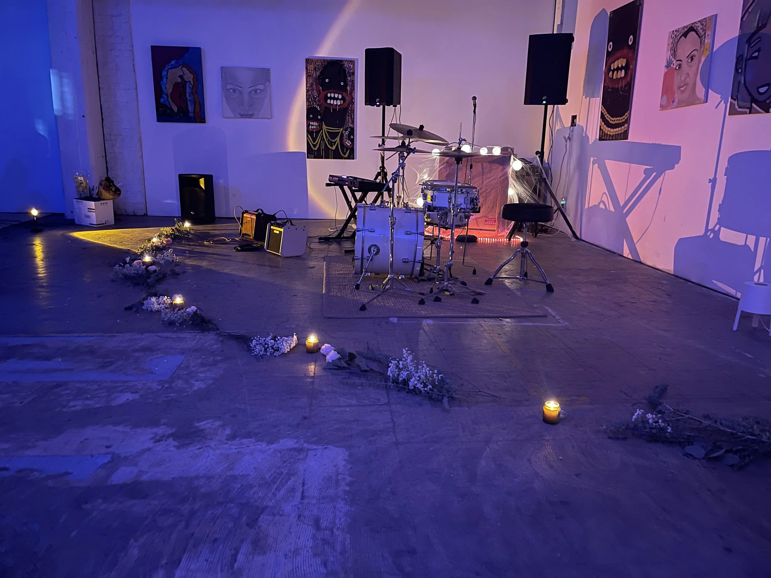 Empty stage set up for a musical performance with a drum kit, keyboard, and speakers, decorated with flowers and candles, illuminated with soft purple and pink lighting.