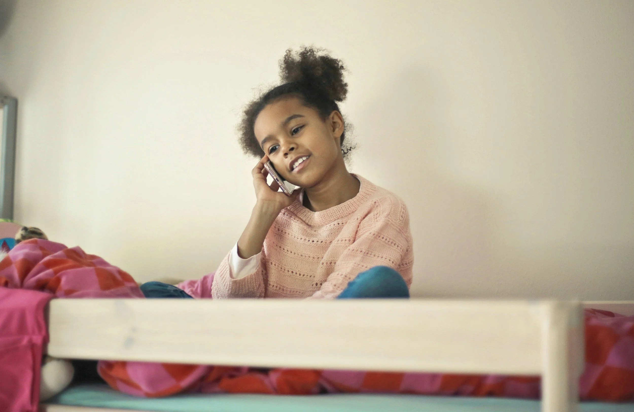 A young girl with curly hair in a messy bun talking on a mobile phone while sitting on her bed.