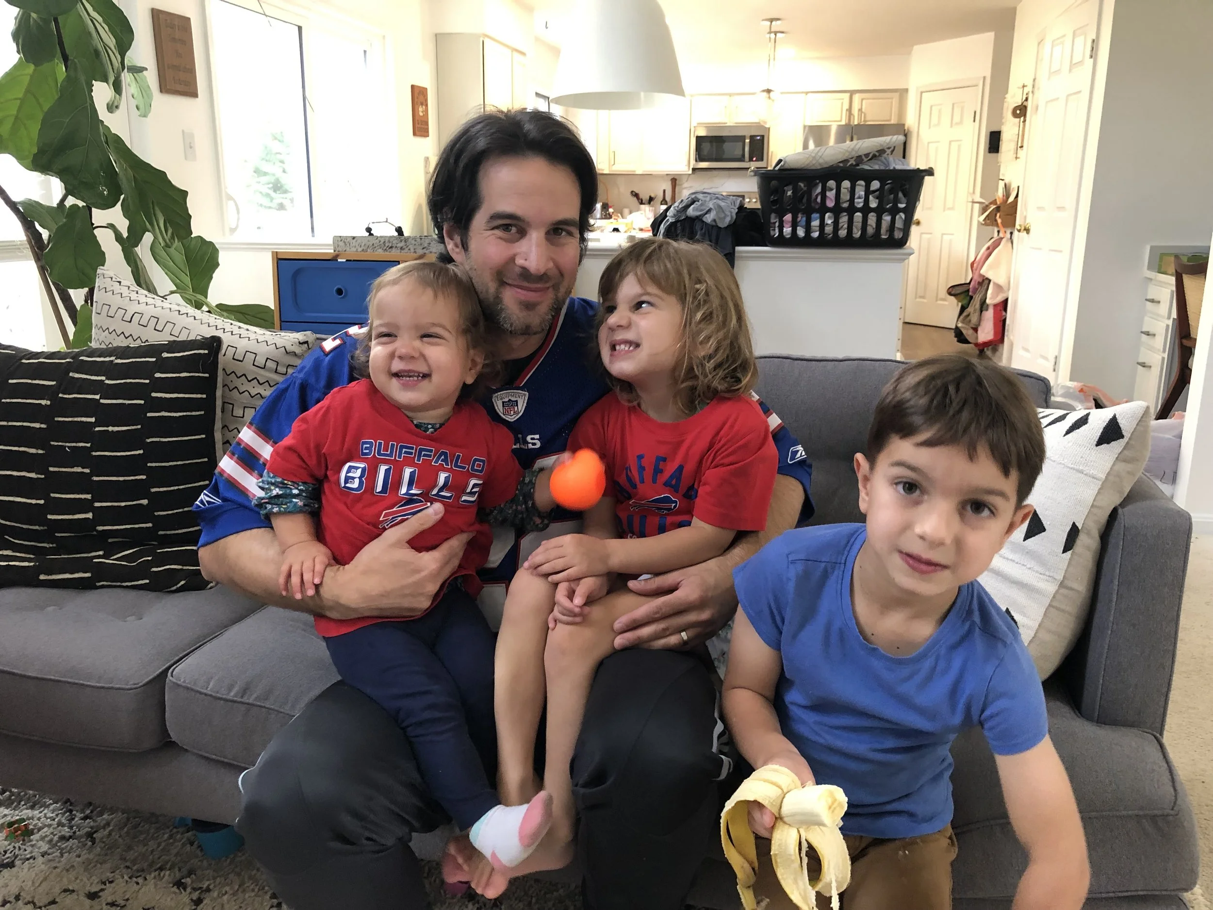 A man in a middle of a living room with three young children, two girls and one boy. The children are smiling and holding snacks, with the girl on the left holding an orange snack, the girl in the middle smiling and looking at the man, and the boy on the right holding a bunch of bananas. The background shows an open kitchen area, a blue cabinet, and some laundry in a black basket.