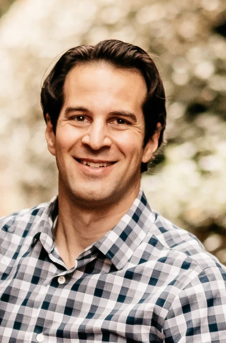 Smiling man with dark brown hair wearing a plaid button-up shirt, outdoors, blurred background.