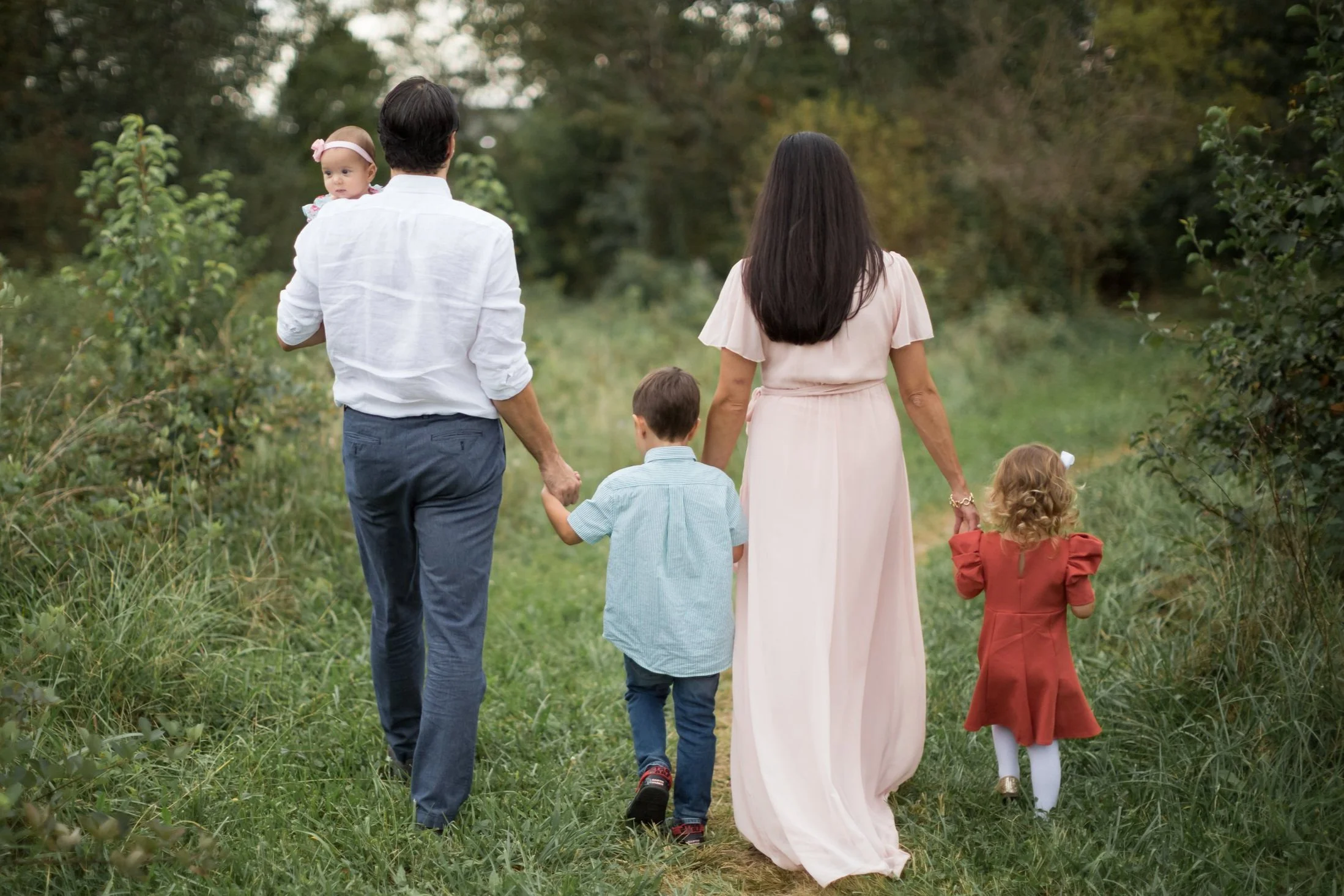 A family of five walking along a grassy, wooded path seen from behind. The father holds a baby girl in his left arm, and the mother, dressed in a flowing pink dress, walks with her hand in her daughter's. Two young children, a boy and a girl, are walking between them. The scene is peaceful and surrounded by trees.