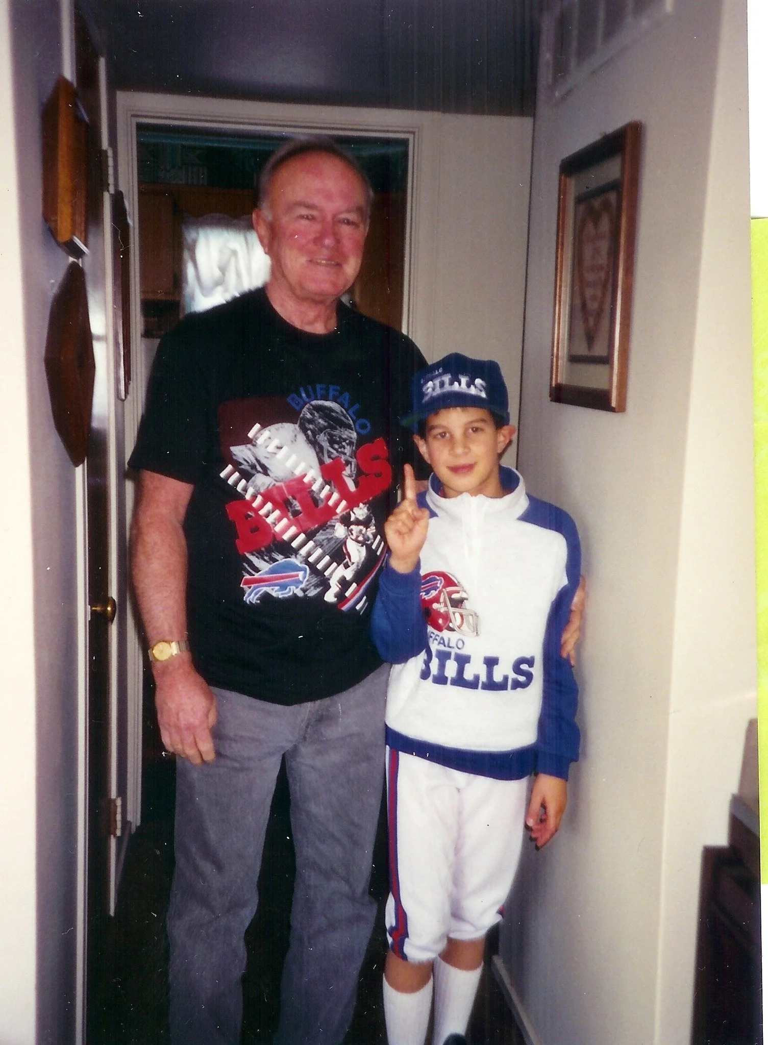 An elderly man and a young boy pose together in a hallway, both dressed in Buffalo Bills fan gear. The man is wearing a black Buffalo Bills T-shirt, while the boy sports a white and blue uniform with the Bills logo and a cap. The man is smiling, and the boy is holding up one finger, possibly indicating the number one.