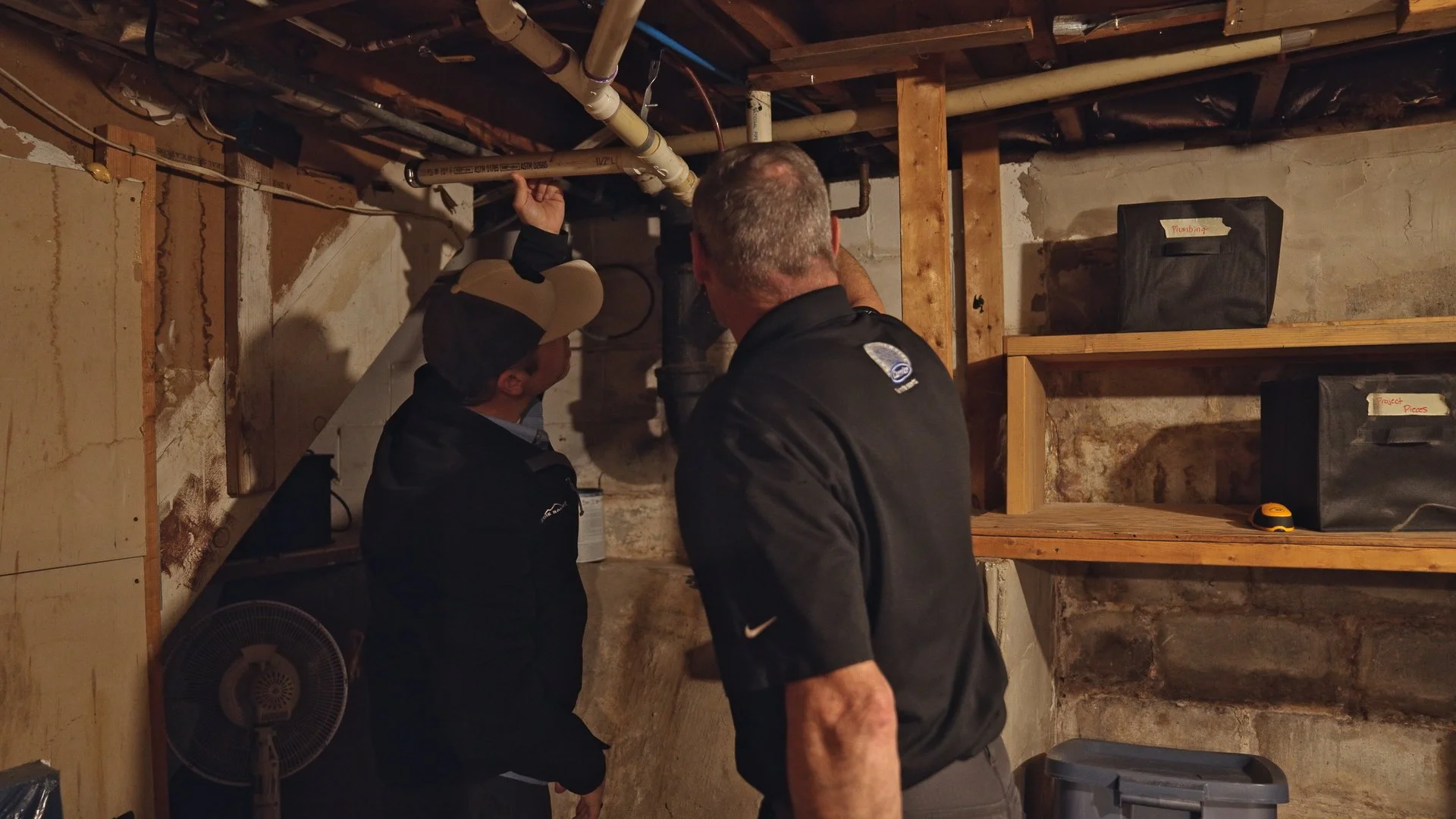Two men looking at pipes in an unfinished basement