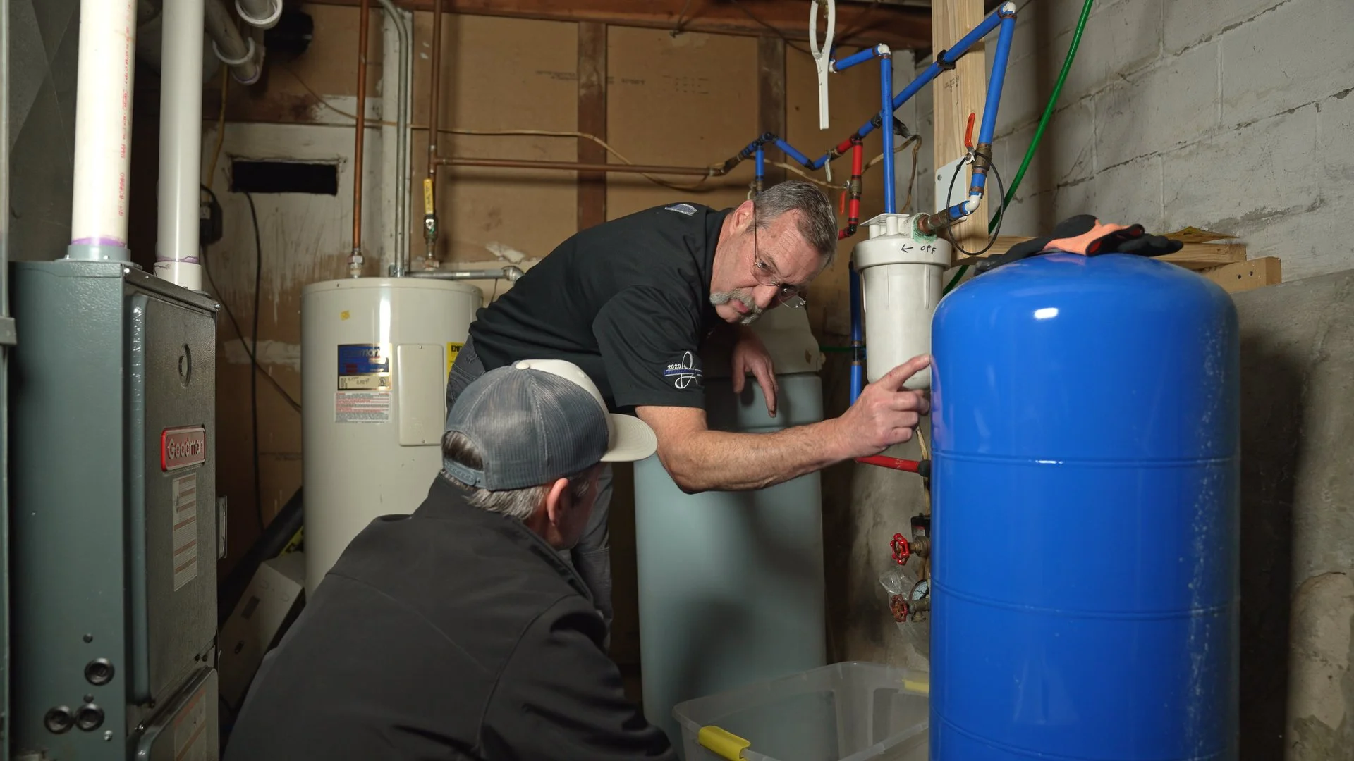 One man pointing out a blue water tank to another man in a hat in an unfinished basement