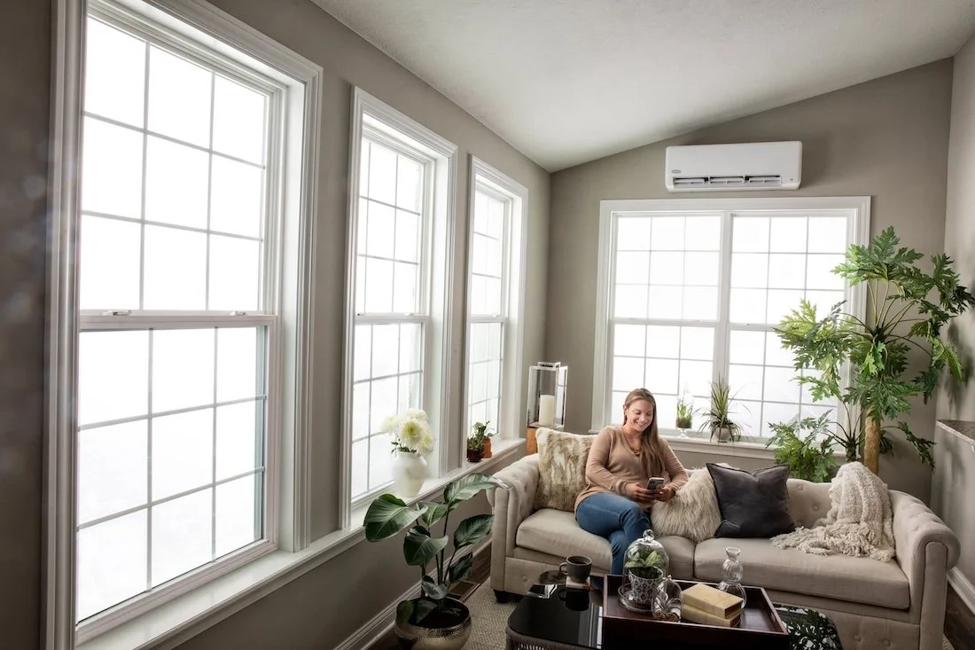 A woman sitting on a beige couch in a living room with large windows, a potted plant, and a coffee table.