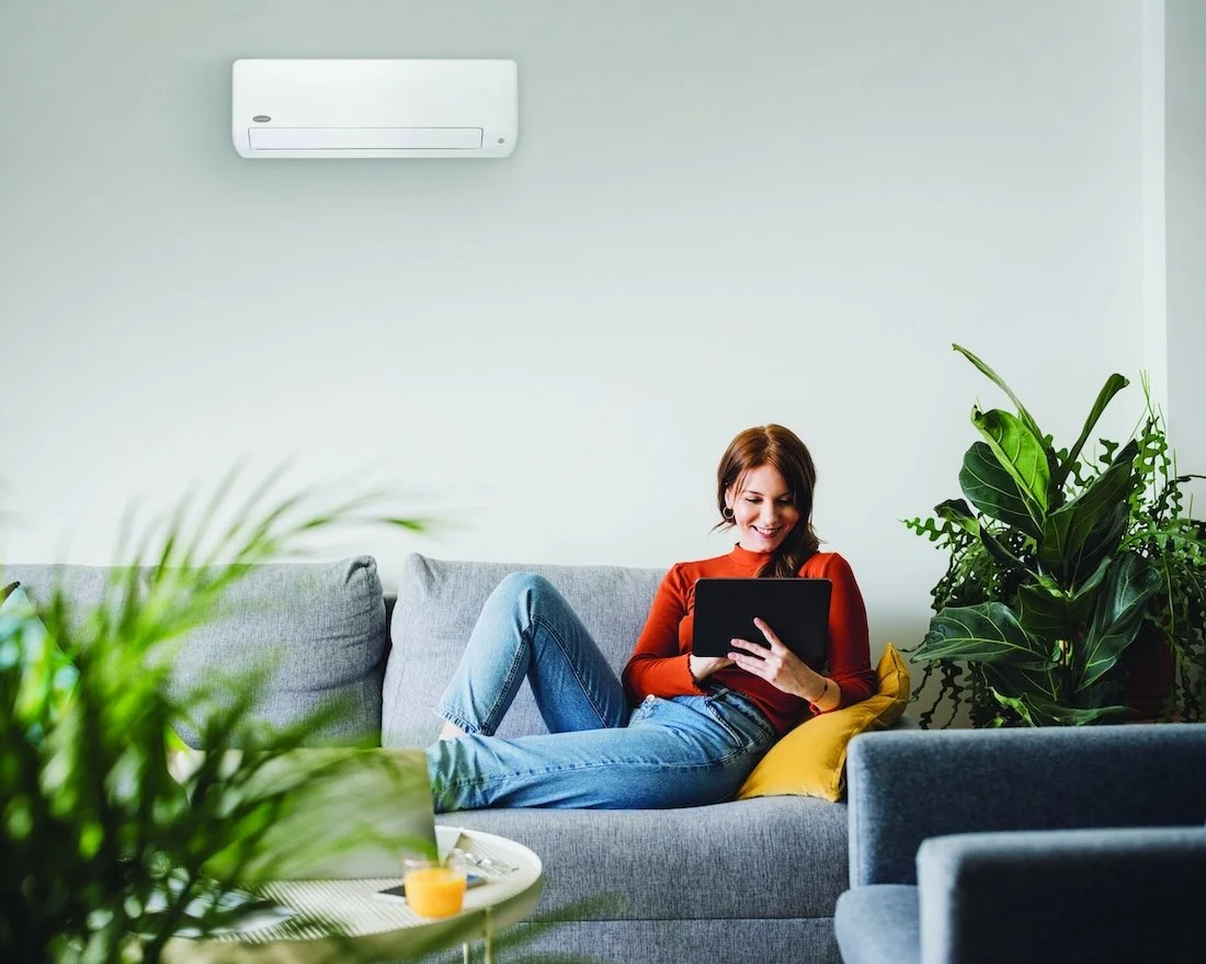 A woman with red hair sitting on a light gray couch, reading a tablet, surrounded by green houseplants in a bright, modern living room with a wall-mounted air conditioner.