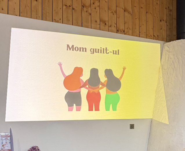 A projected slide on a wall with an illustration of three women with different hair colors and styles holding hands, under the text "Mom guilt-ul" in brown font.