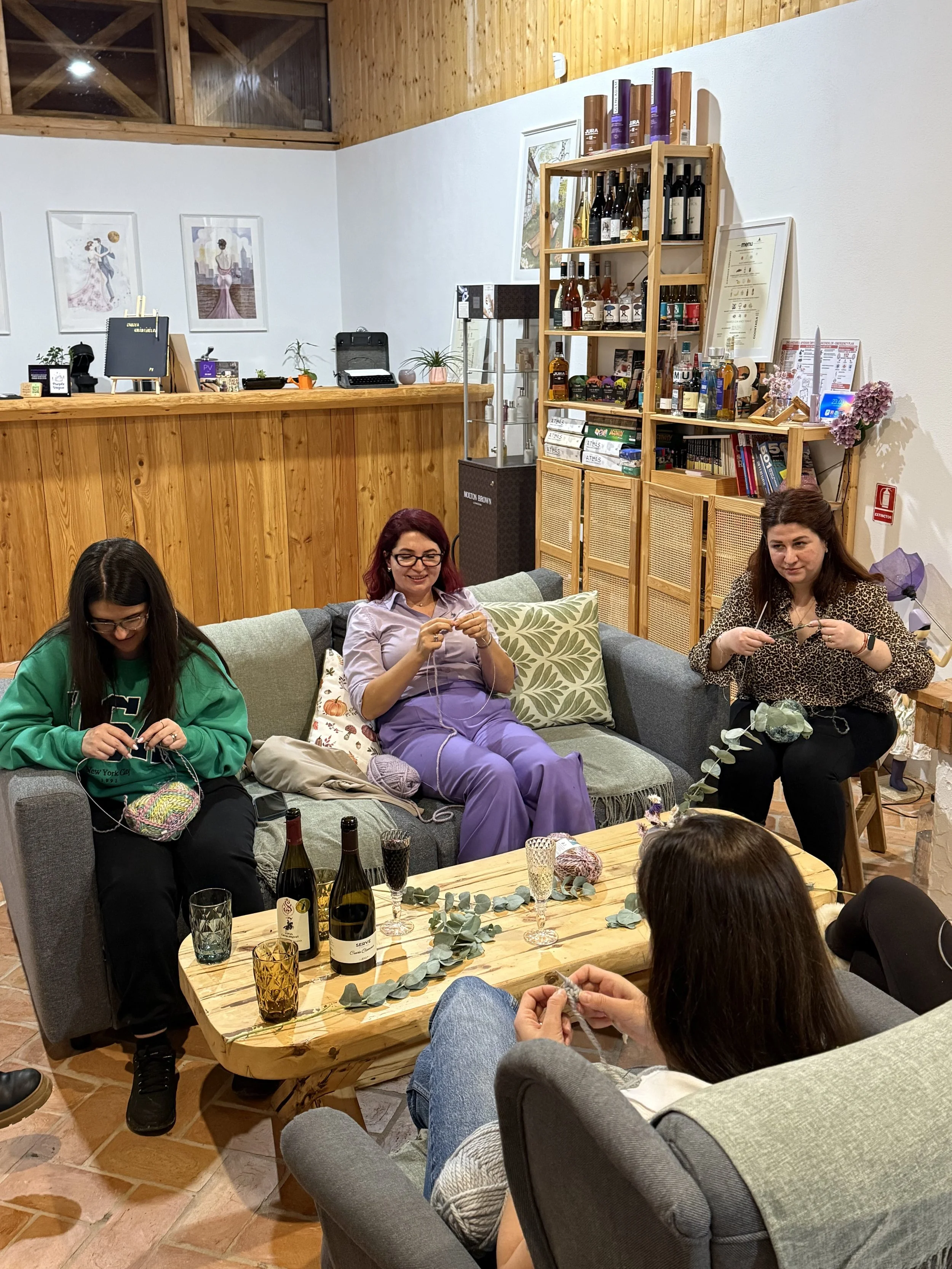 Four women sitting in a cozy cafe knitting and socializing around a wooden table with wine bottles and glasses, with a bar and decorative art on the wall in the background.