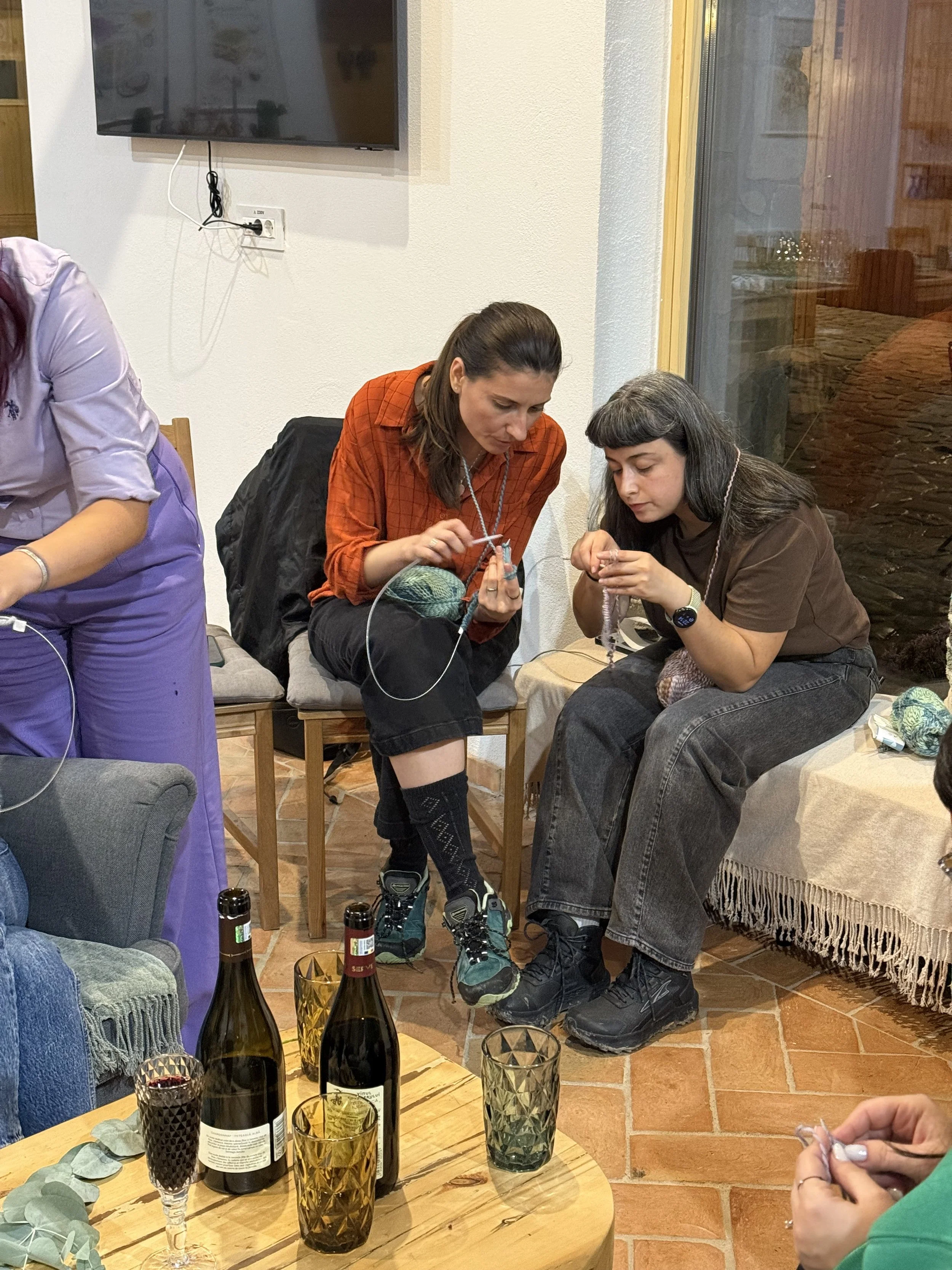 Two women sit on chairs knitting, with bottles and glasses on a table in front of them, in a cozy indoor setting.