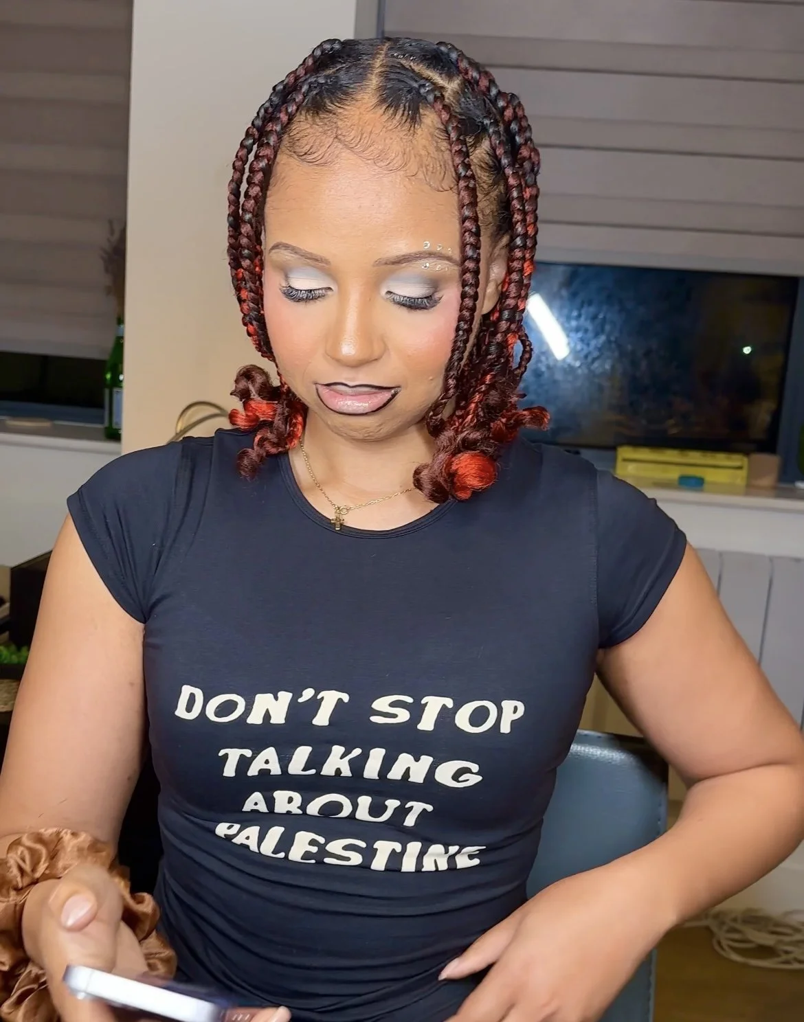 Woman with styled braids wearing a black T-shirt that says 'DON'T STOP TALKING ABOUT PALESTINE' in white lettering, sitting indoors, looking down, with long eyelashes and makeup.