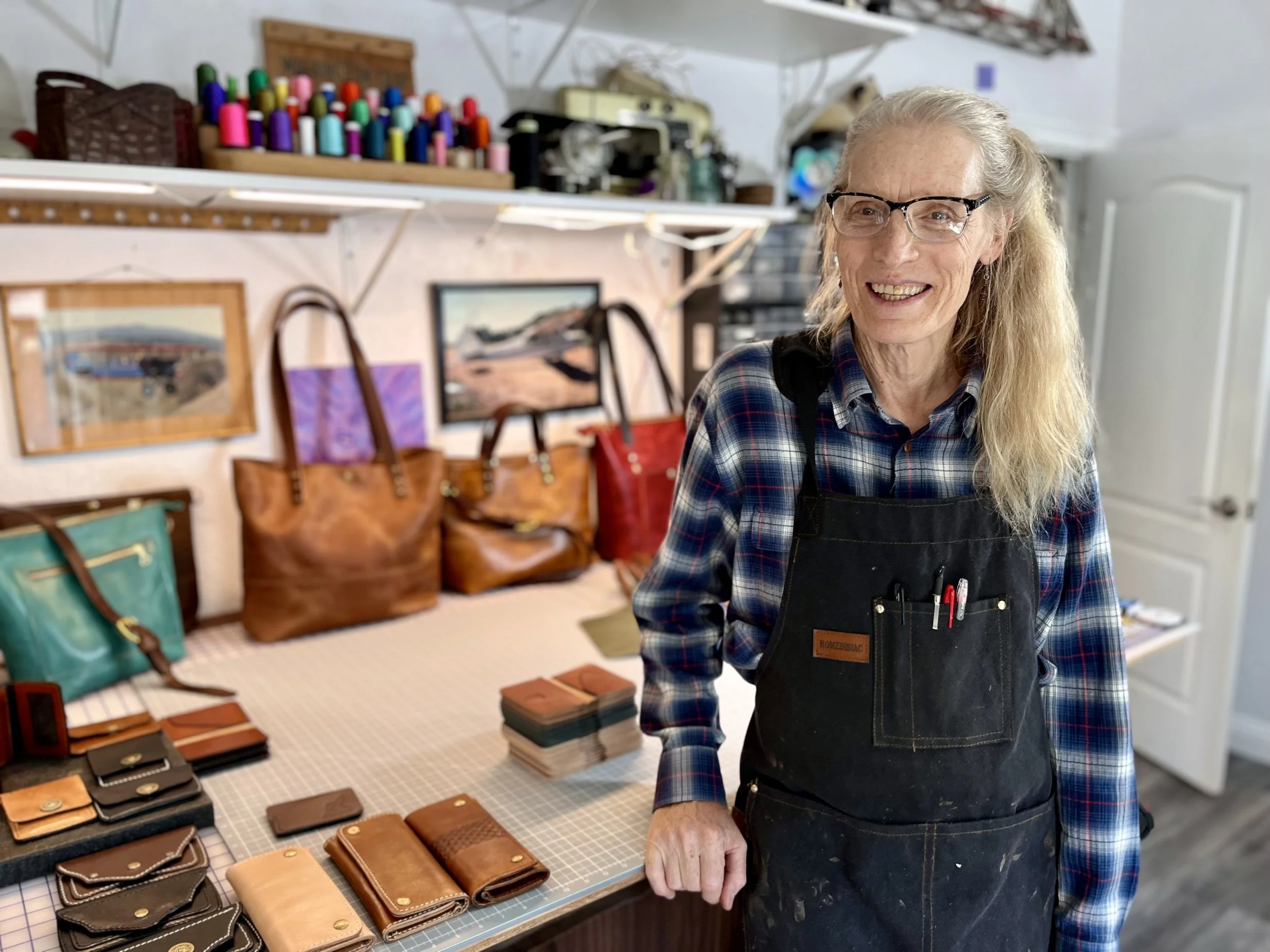 Jamie from Howling Leathers standing inside the Howling Leathers workspace with a workstation holding several handcrafted leather wallets and bags.