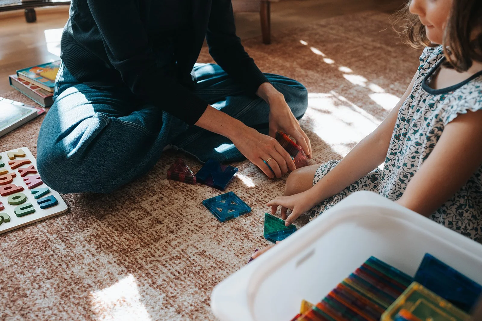 A woman and a girl playing with colorful magnetic building tiles on a carpeted floor in a room with sunlight streaming through a window.