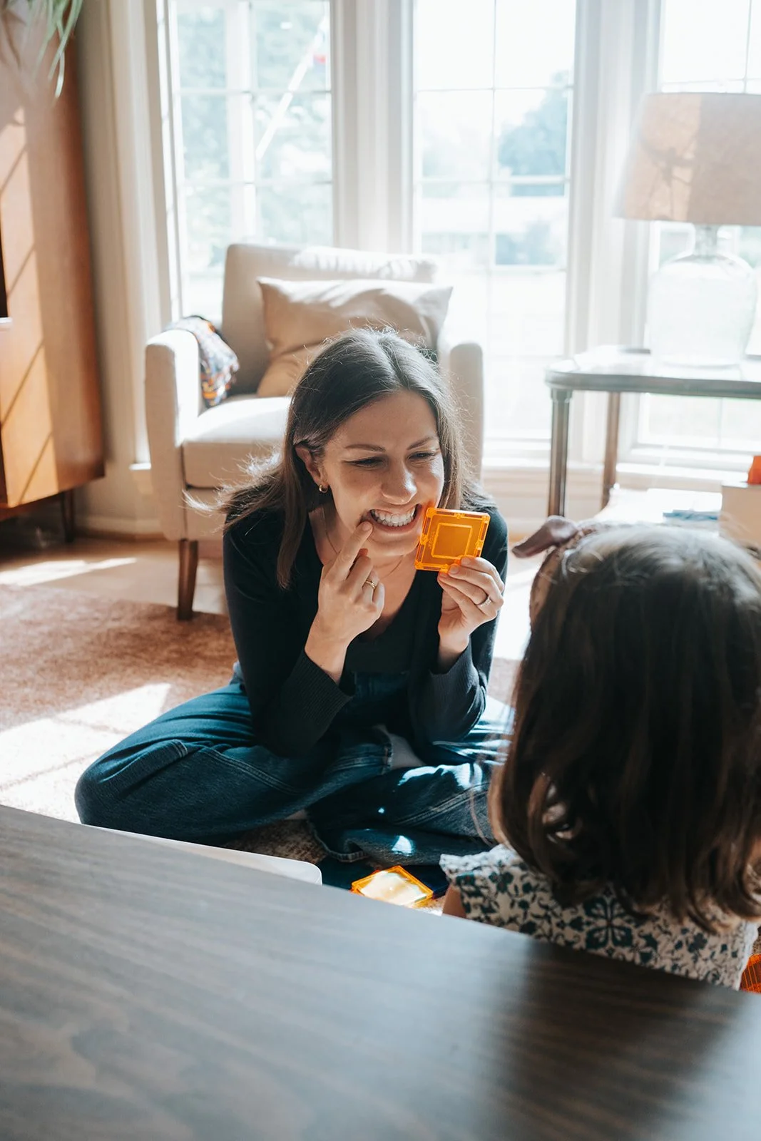 A woman and a young girl playing a card game on the floor of a sunlit living room.