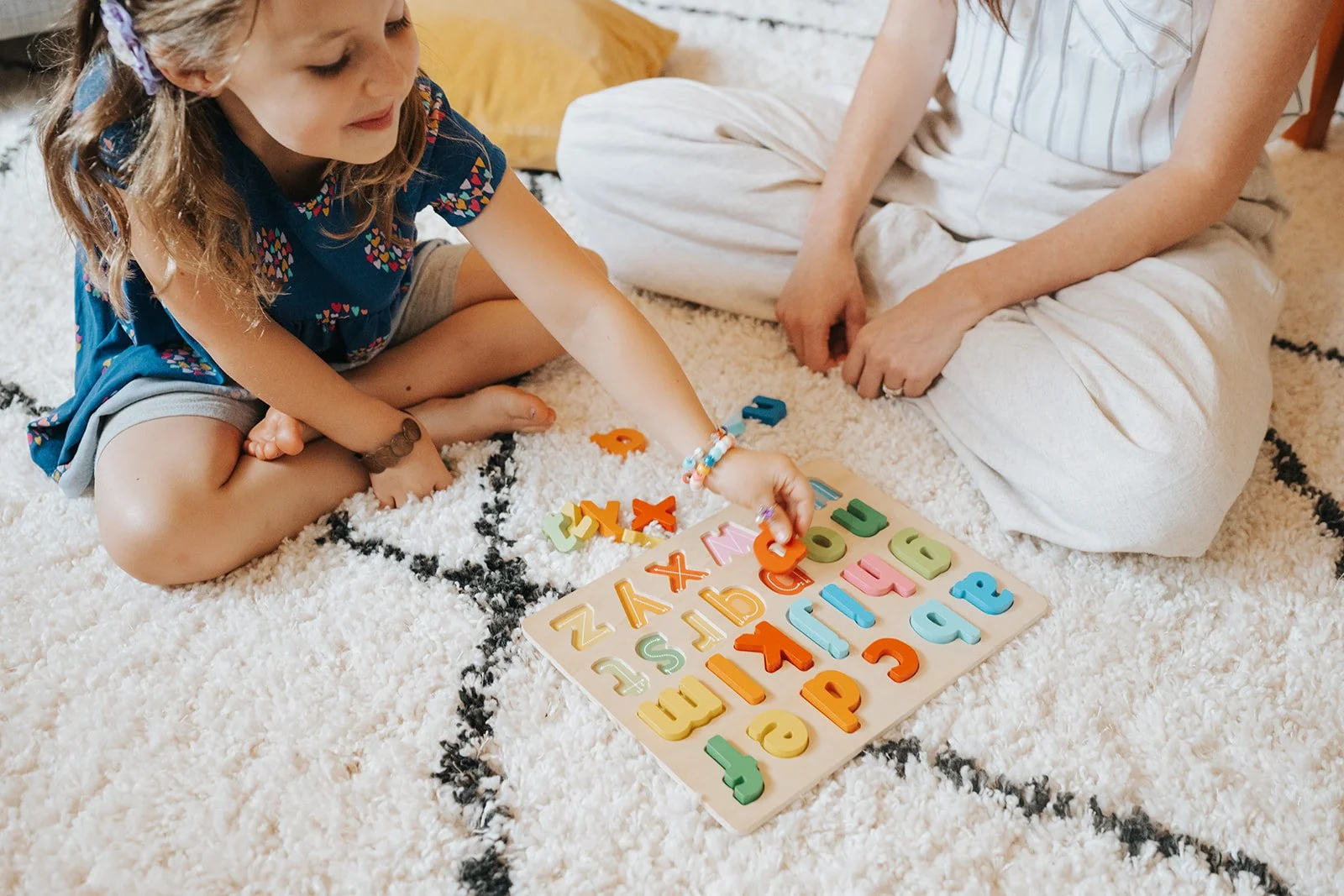 A young girl with a colorful bracelet playing with foam alphabet letters on a wooden board, while an adult sits nearby on a plush white rug with a black pattern.