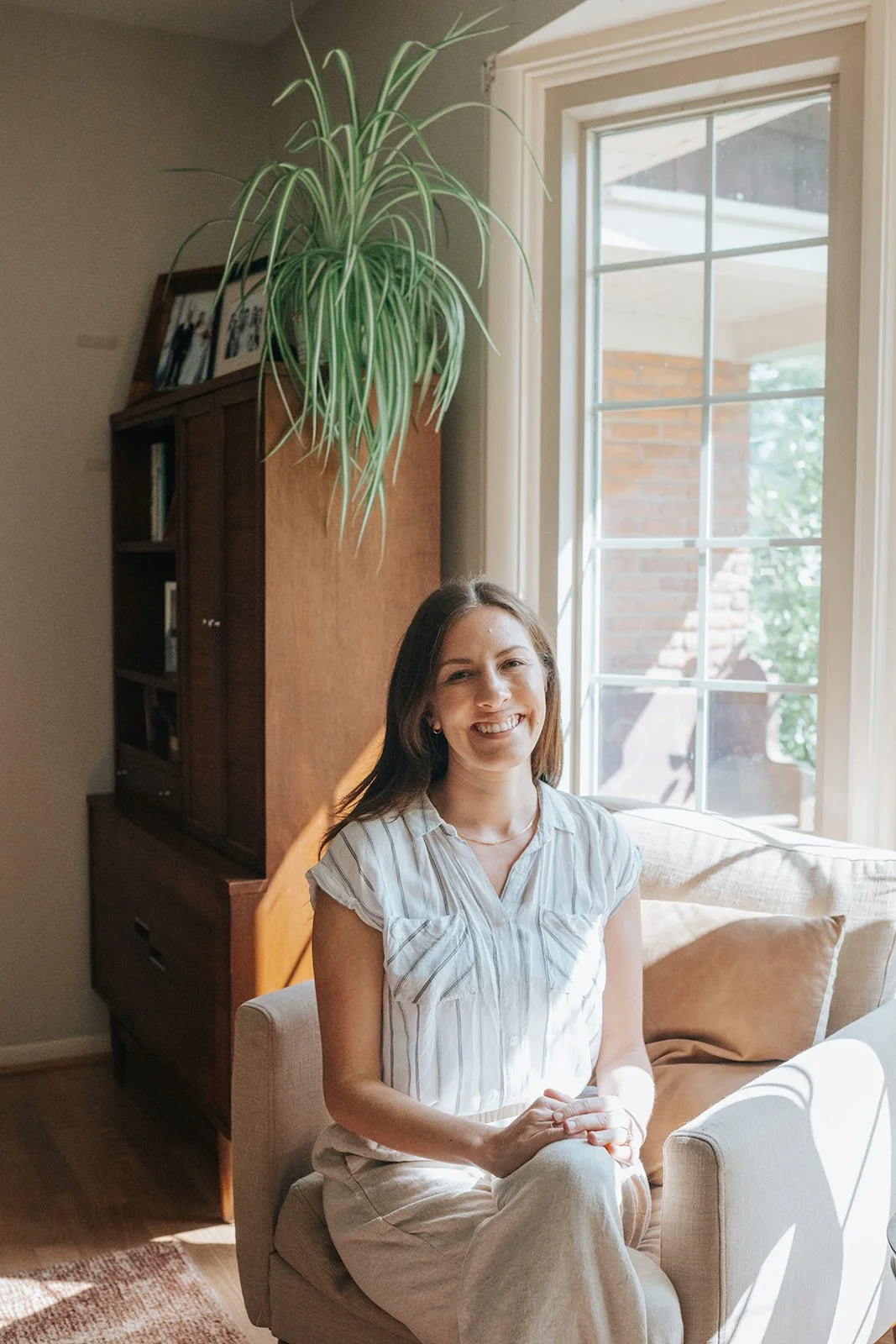 A smiling woman with brown hair sitting on a beige armchair in a sunlit living room next to a window with white trim, a wooden cabinet, and a hanging green plant.