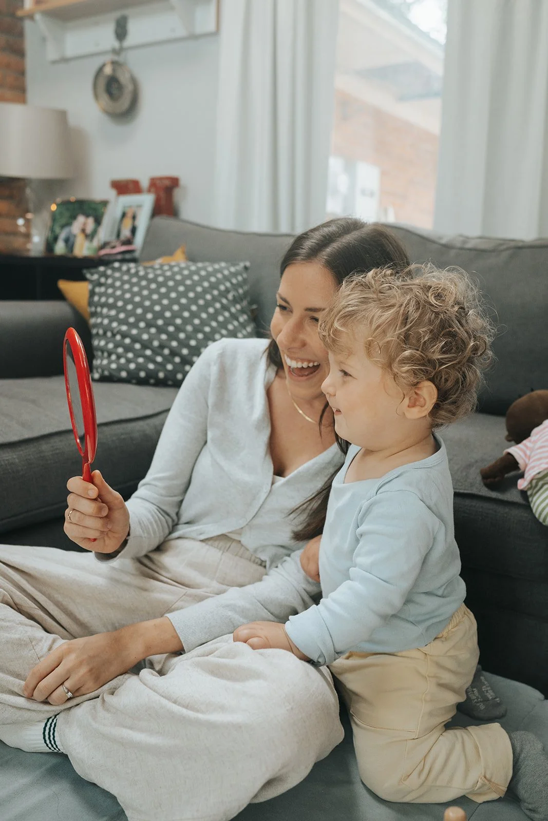 A woman and a young child sitting on the floor, looking at a red handheld mirror together and smiling. They are in a cozy living room with a gray sofa, decorative pillows, and personal photos on a side table.