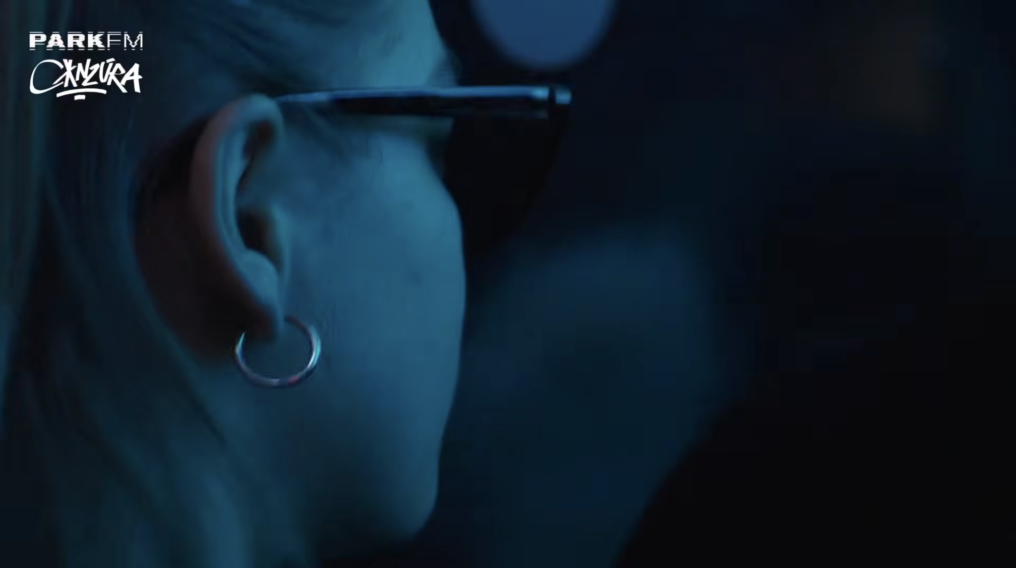 Close-up of a person with glasses and an earring, looking to the side, dark background.