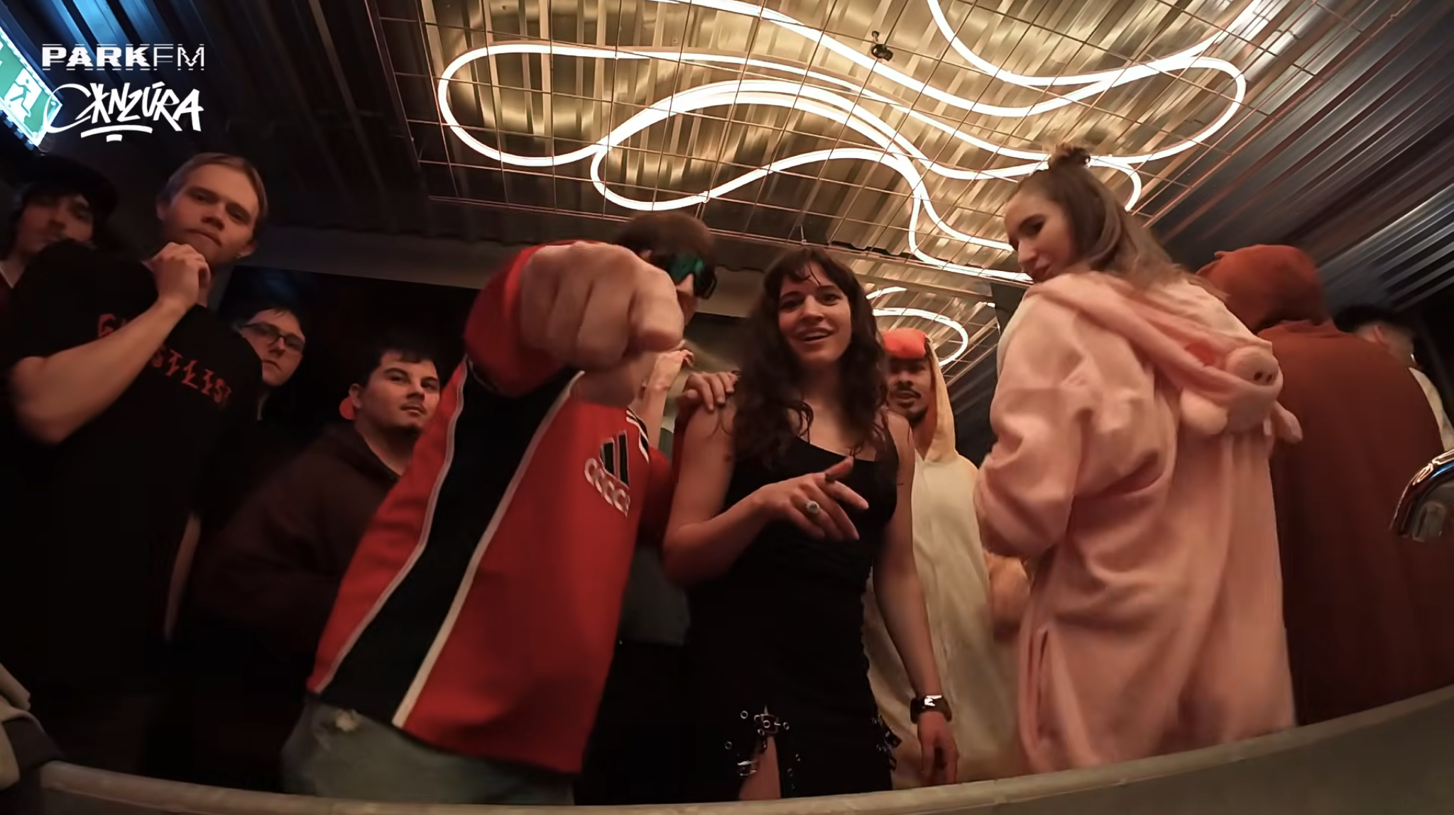 Group of young people indoors, some dressed in costumes, with a modern ceiling light design above, at a social event or party.