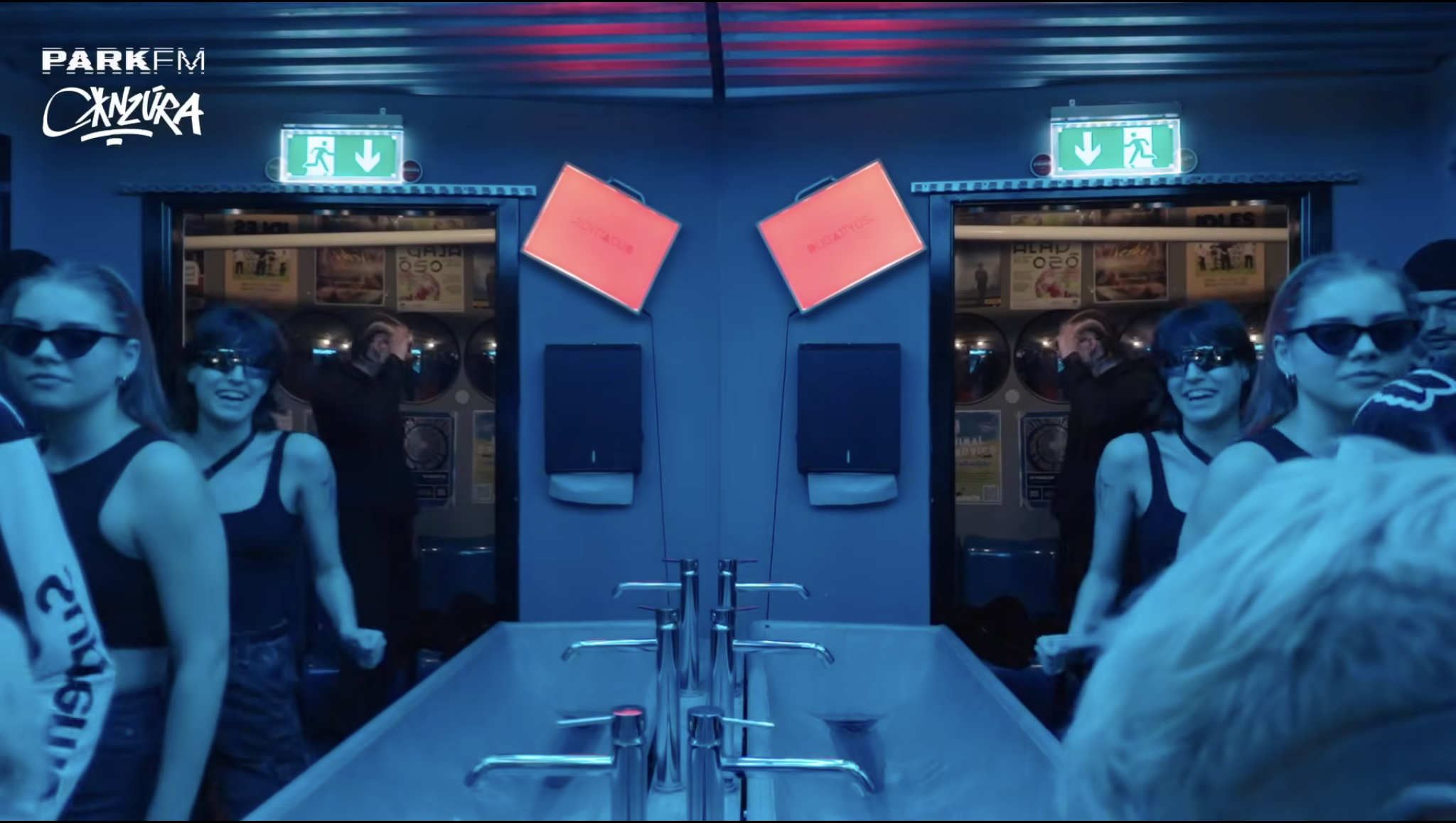 A group of women wearing sunglasses and sportswear standing at a bar counter in a dimly lit, blue-toned environment with neon exit signs overhead and posters on the walls.