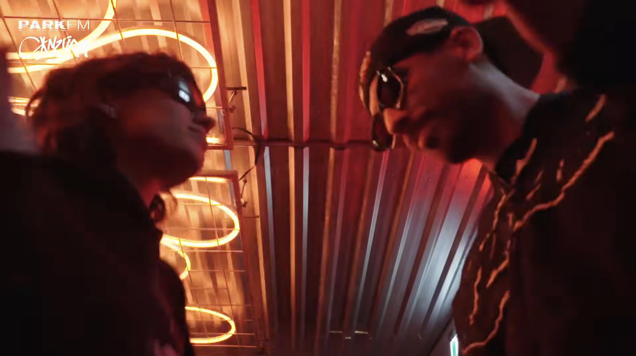Two men wearing protective goggles in a dimly lit room with wooden walls and neon neon signs. The lighting is red and orange, contributing to an ambient atmosphere.