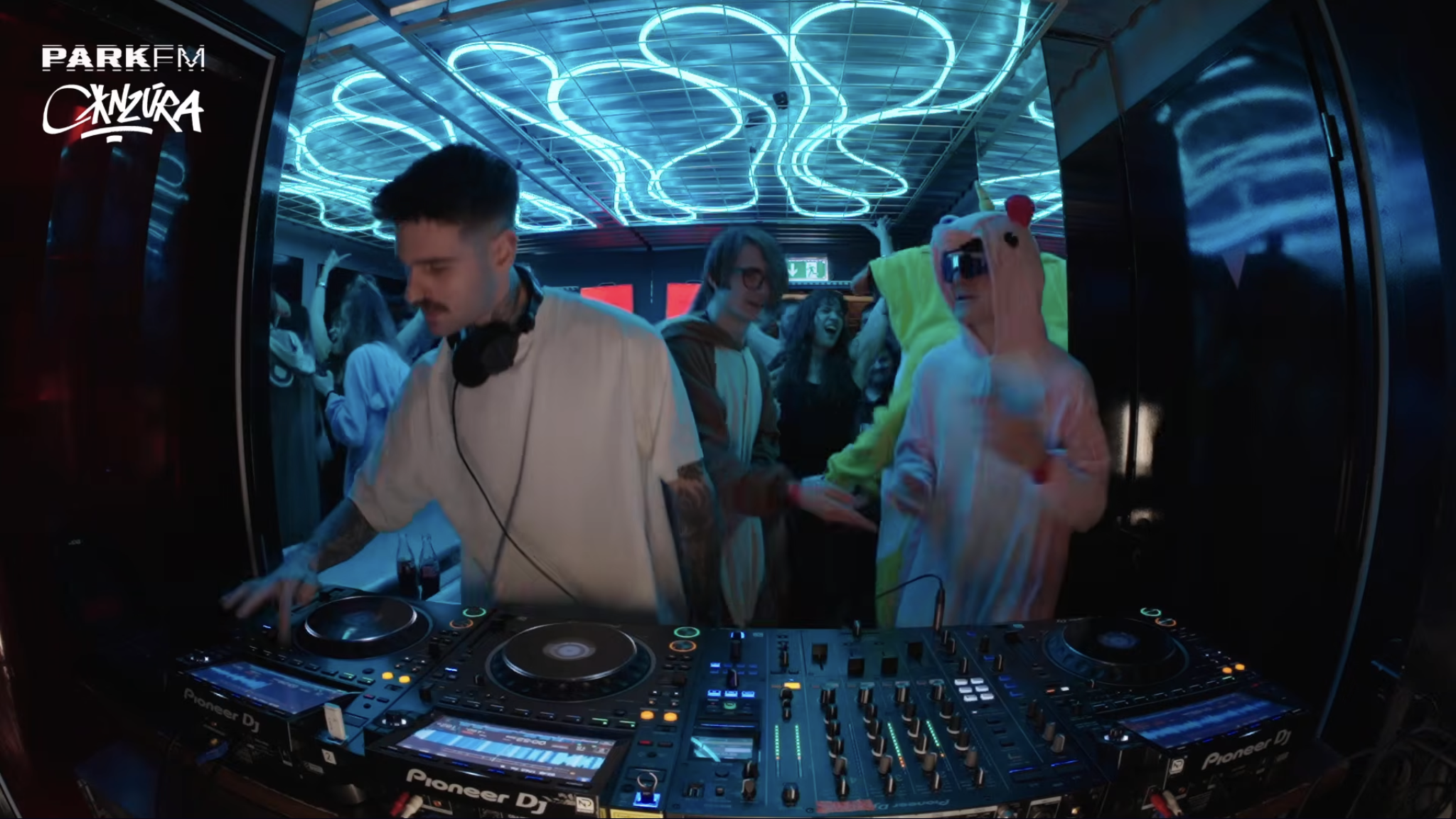 A DJ is playing music at a nightclub with a neon blue ceiling and a crowd of people dancing, some wearing costumes, including a person in a pink unicorn hoodie.