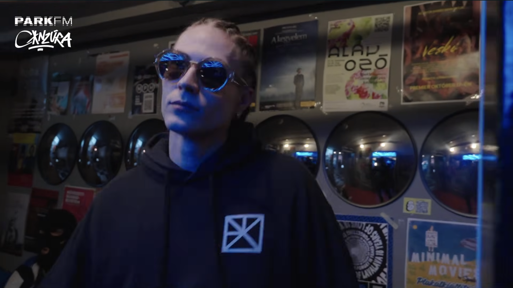 A person wearing sunglasses and a black hoodie with a geometric design stands in front of a vending machine with posters, advertisements, and stickers on the wall behind him.