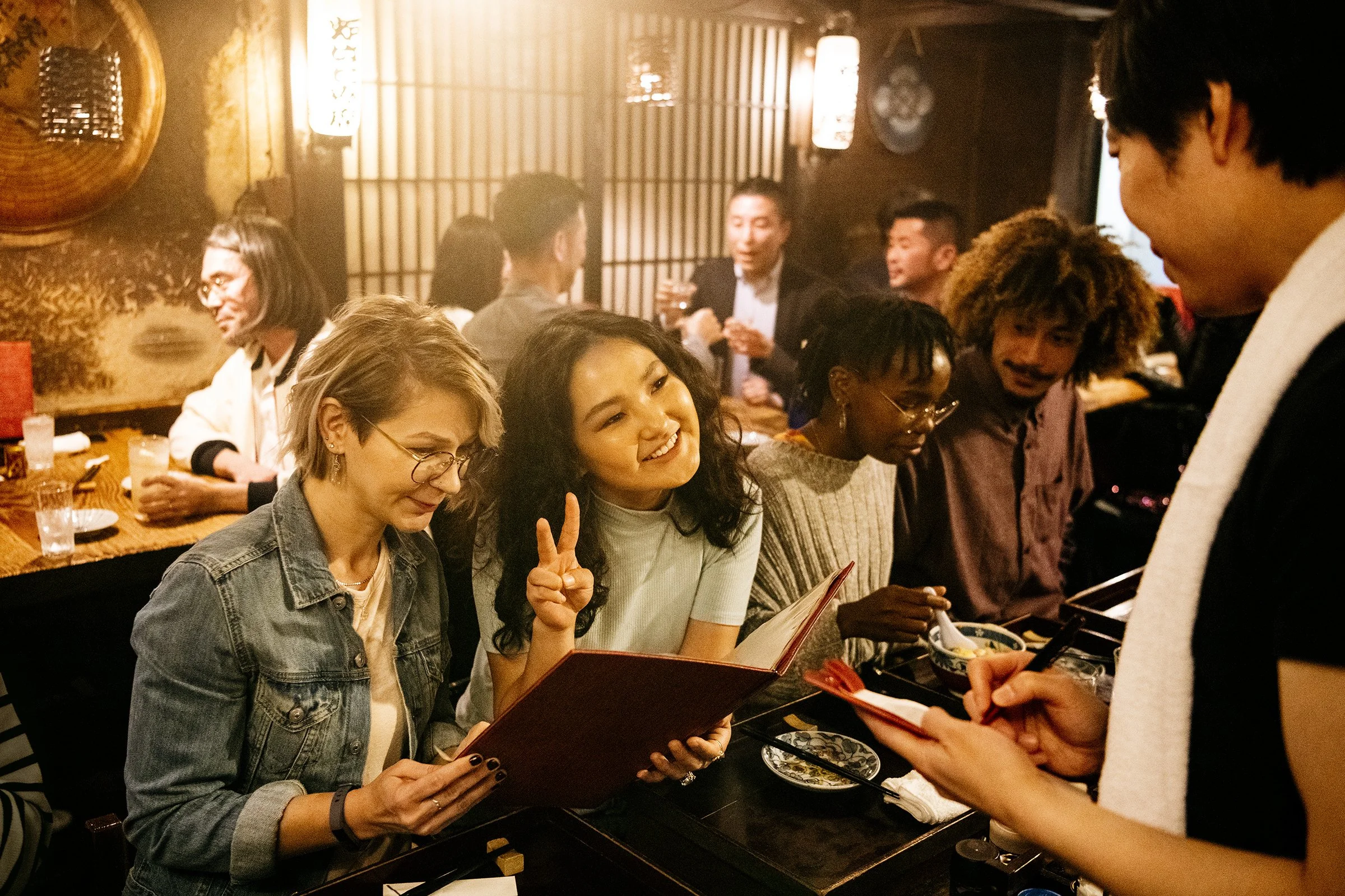 Group of people in a cozy restaurant, one person holding a menu, others enjoying food and drinks, dimly lit atmosphere.