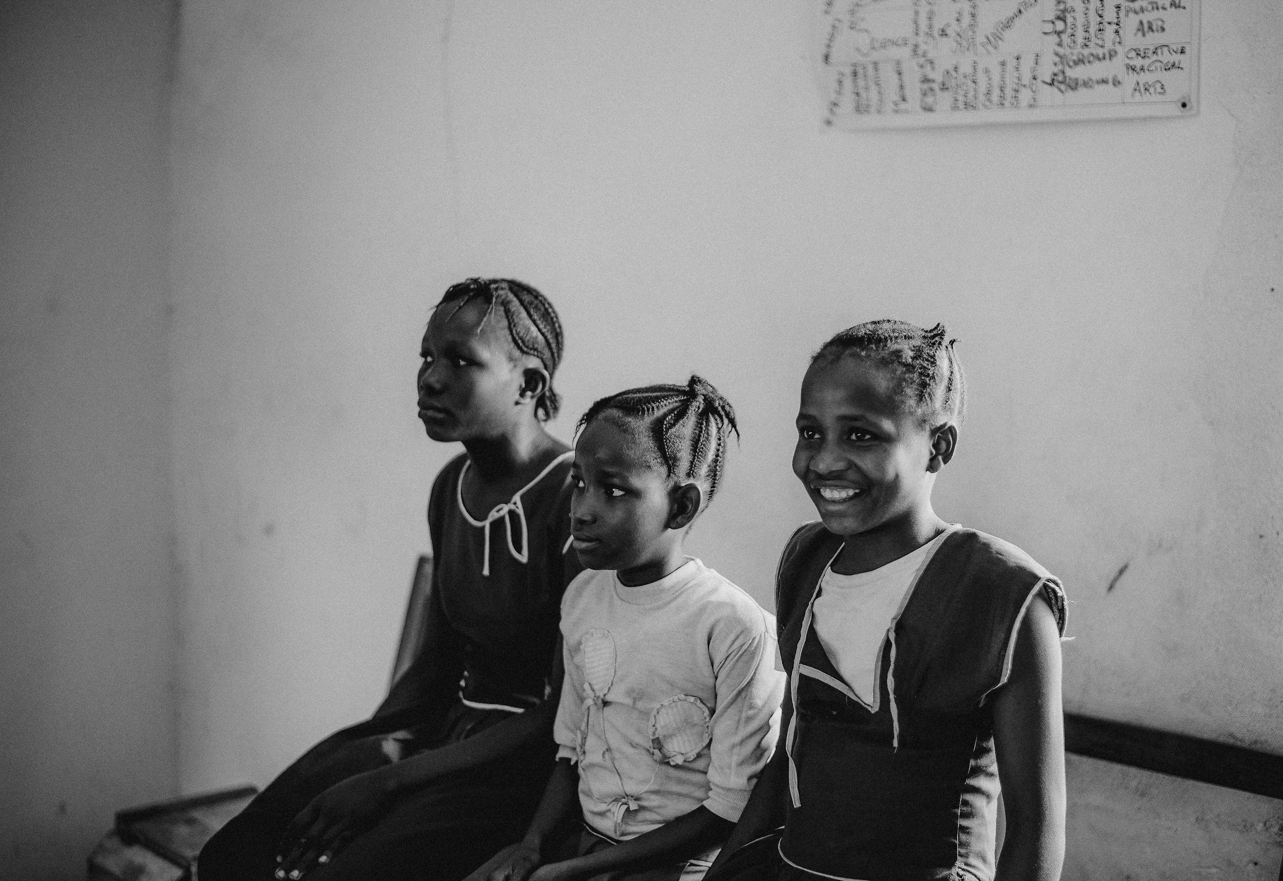 Three young girls sitting on a bench against a plain wall, one smiling and the others looking away, in a classroom or waiting area.