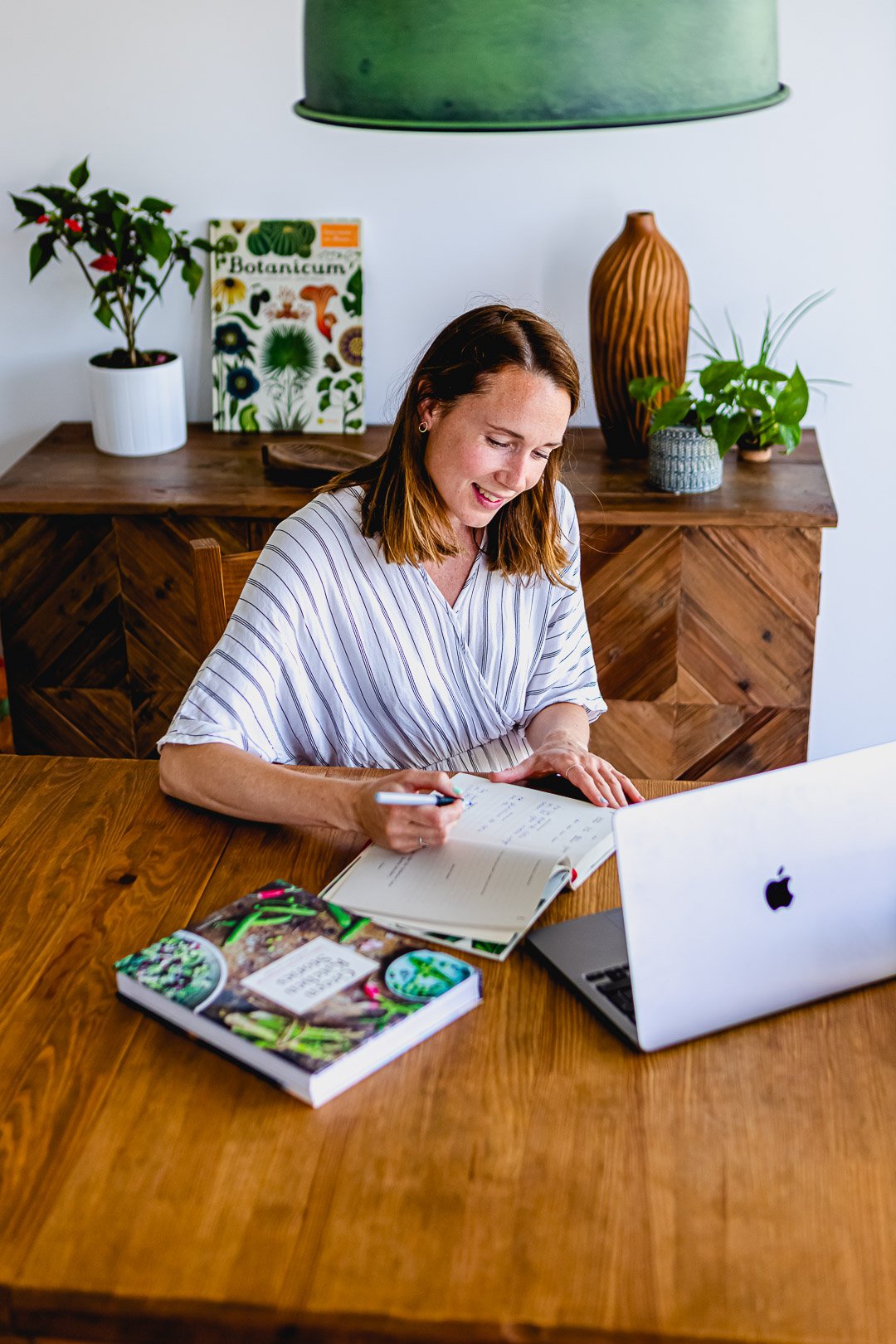 Woman writing in a notebook at a wooden table with a laptop and a botanical book, surrounded by plants and a vase.