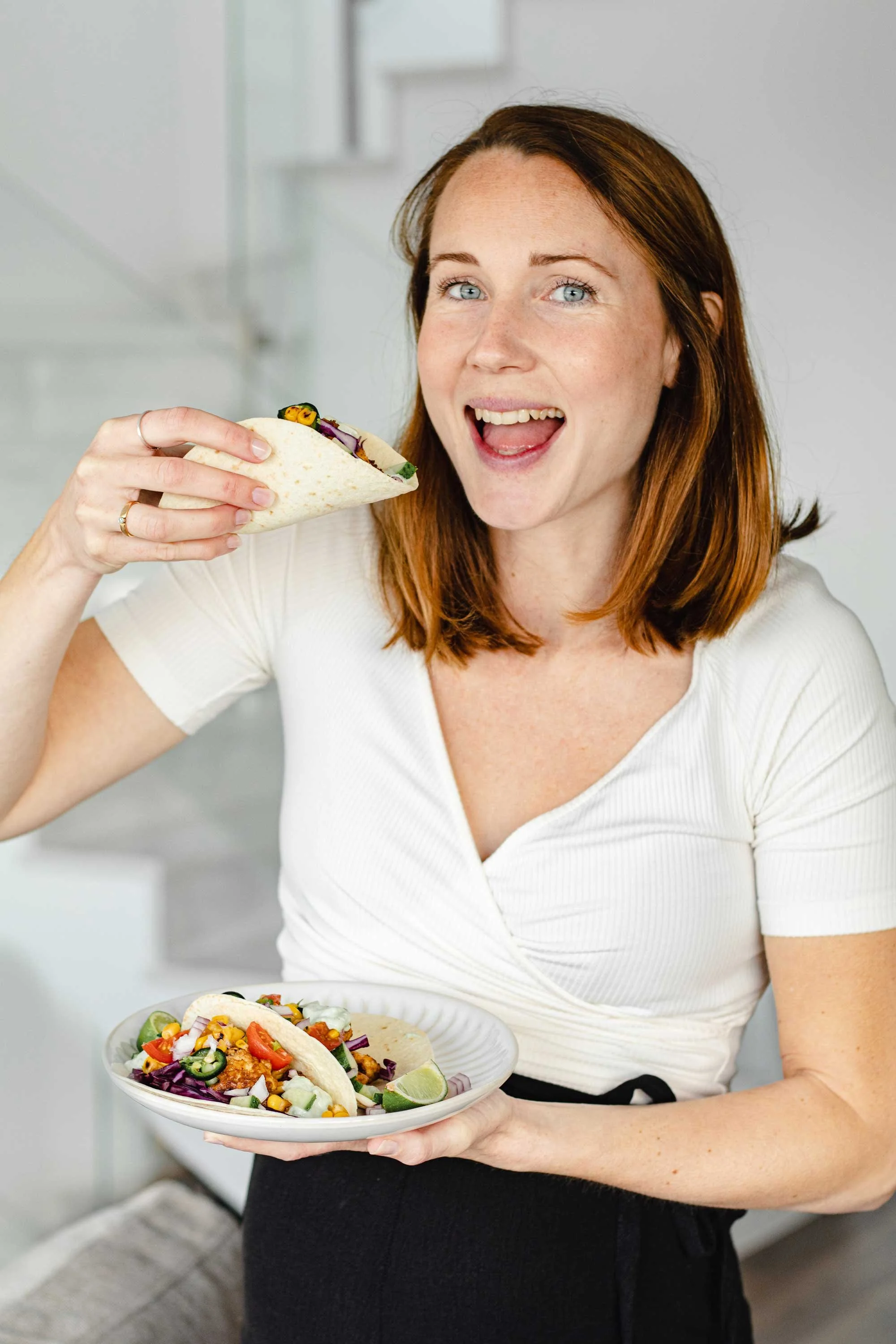 Woman holding and eating a vegetarian taco while smiling, with a plate of tacos in her other hand.