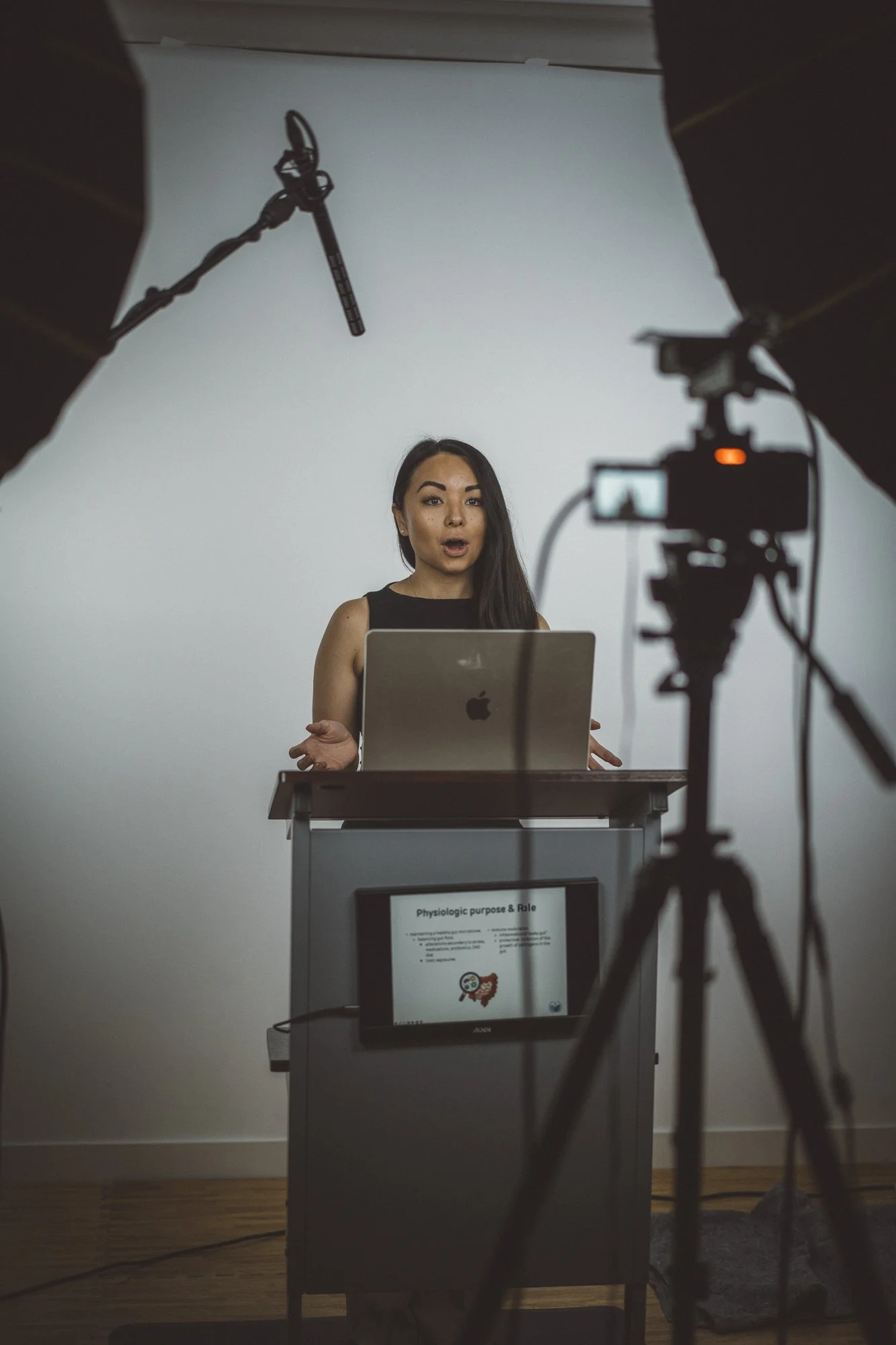 A woman giving a presentation in a studio, standing behind a podium with a laptop and monitor displaying a slide titled 'Physiologic purpose & Role,' with softboxes and camera equipment visible.