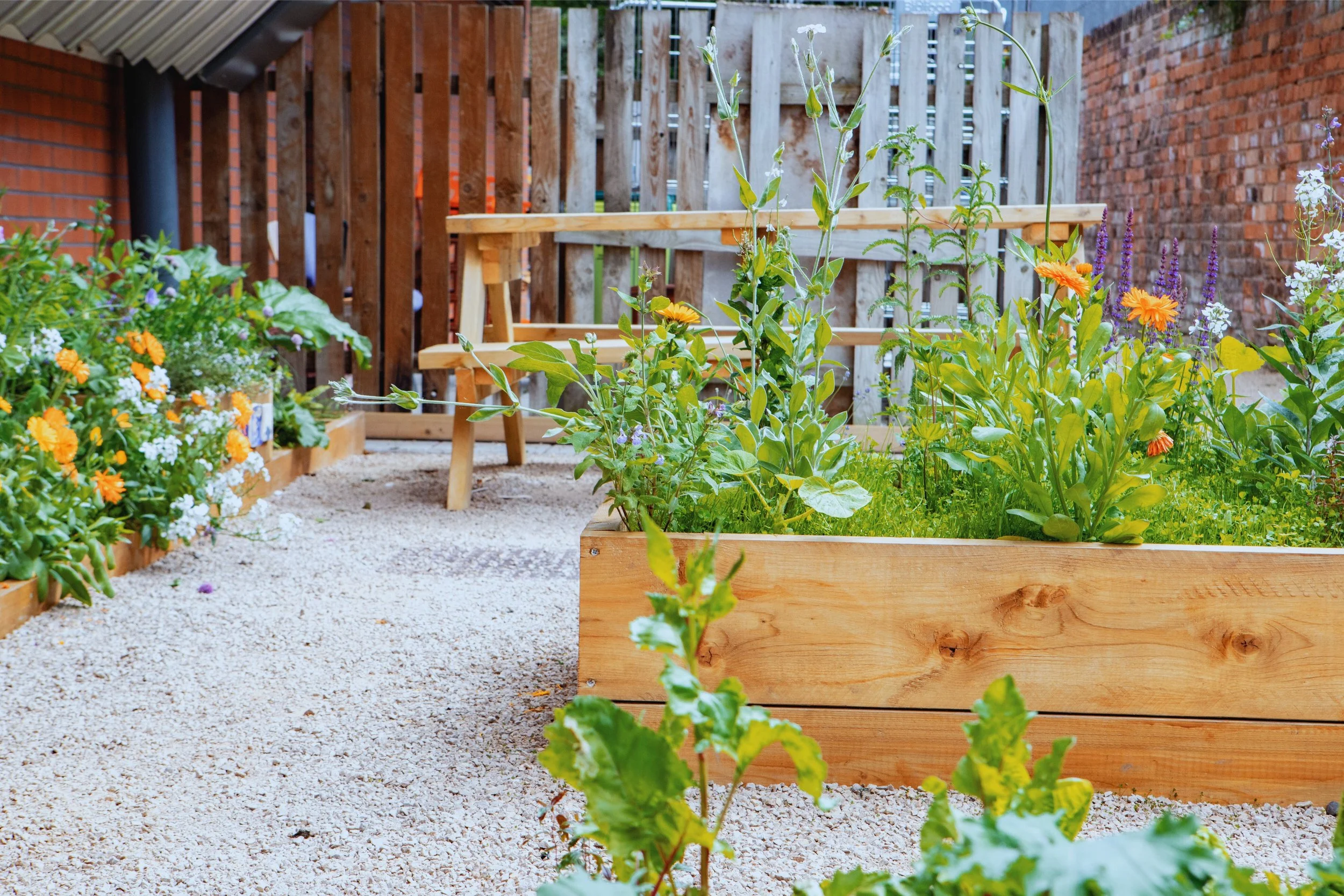 A small garden with wooden planters filled with blooming flowers and green plants, gravel pathway, wooden fence, and a picnic table in the background.