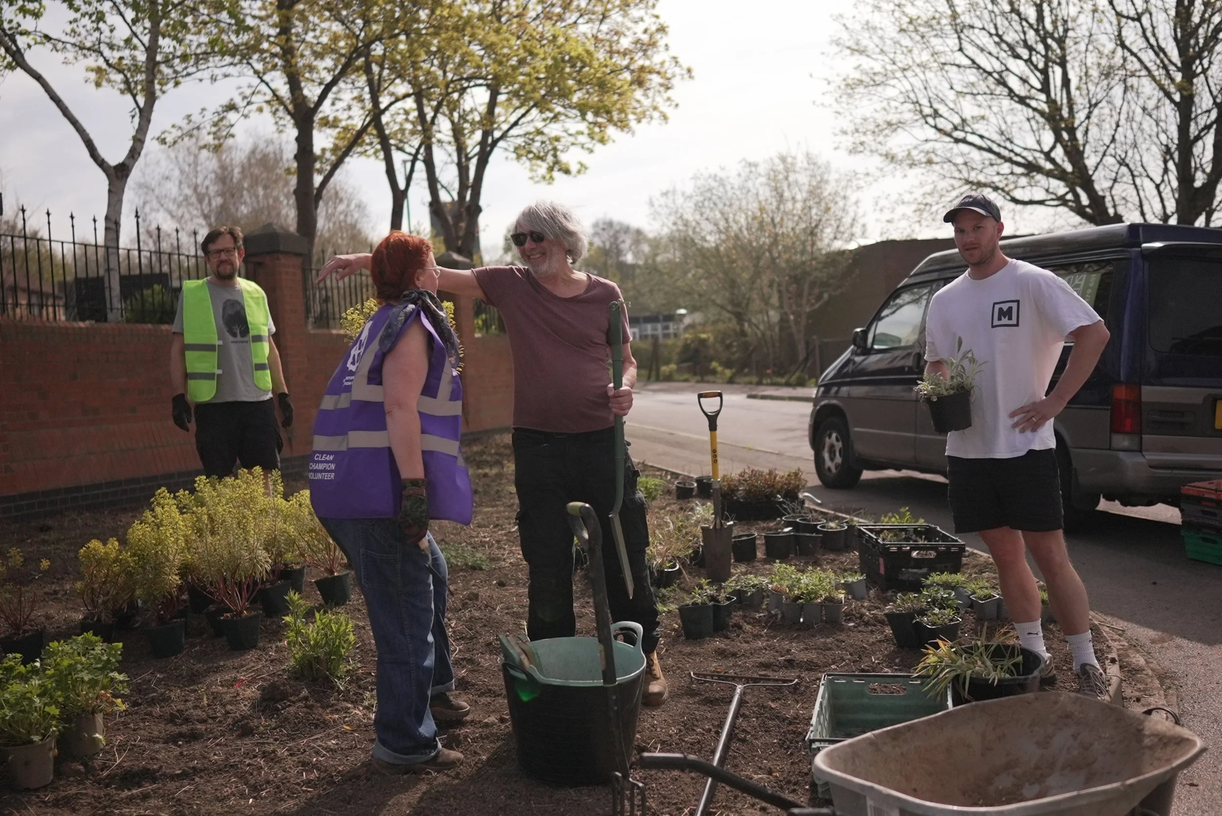 Group of four people planting and tending to a garden on the side of a street, with plants, gardening tools, and a van parked nearby under a partly cloudy sky.
