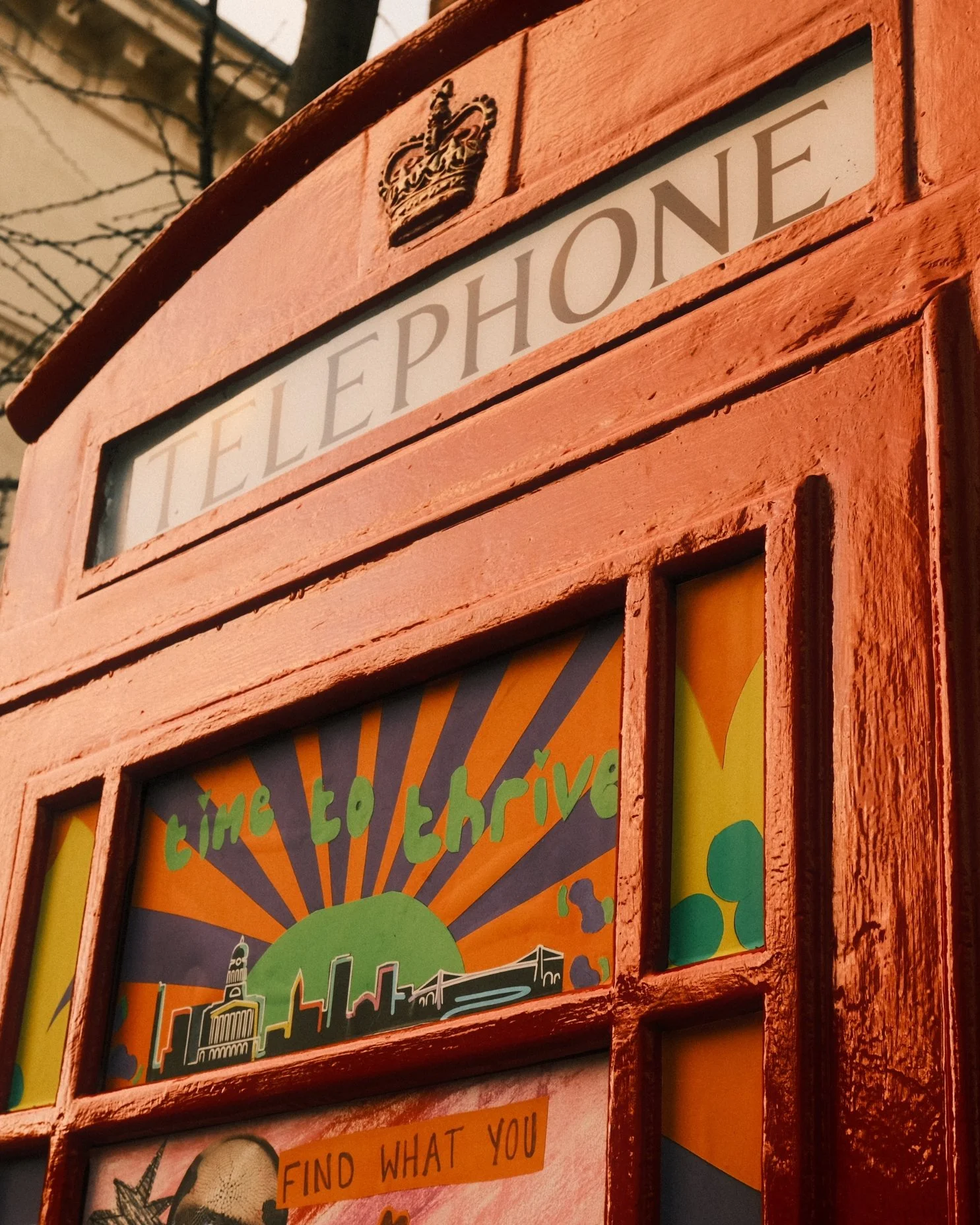 A red British-style telephone booth with the word 'TELEPHONE' and a crown emblem on top. Inside, there's a colorful sign that reads 'Time To Thrive' with a cityscape and sunrise/sunset graphic.