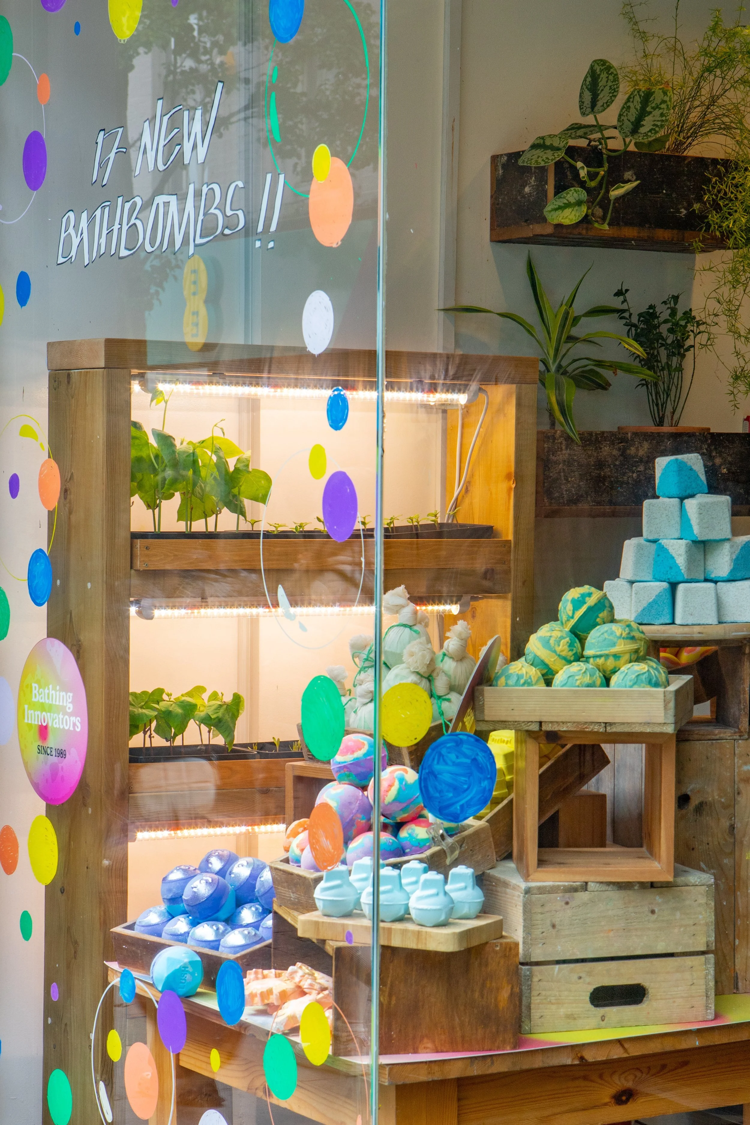 Display window decorated with colorful polka dots and text announcing '17 New Bathbombs!!', showing shelves with bathbombs of various colors and shapes, plants, and wooden storage boxes inside a bath product store.