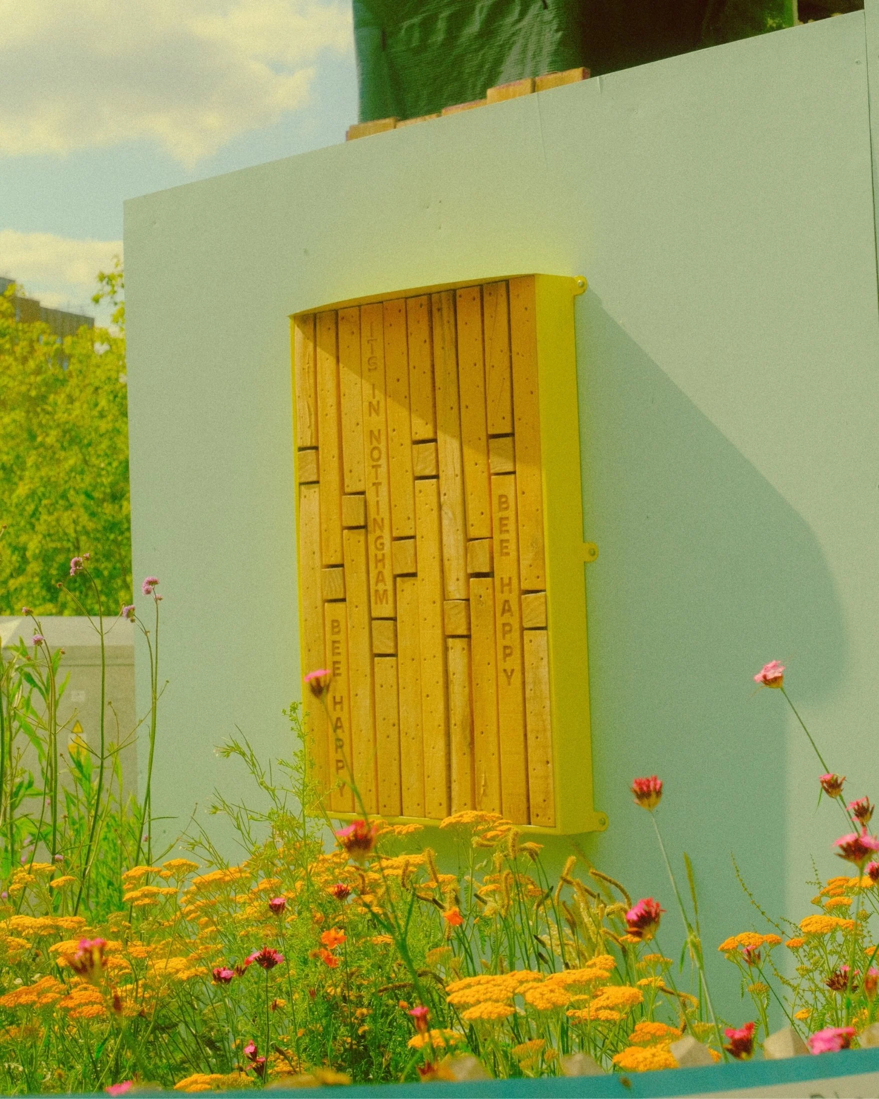 A decorative wall feature with wooden blocks mounted on a turquoise wall, with words like 'BE HAPPY' and 'BEER HAPPY' engraved on the blocks, and surrounded by colorful flowers in the foreground.