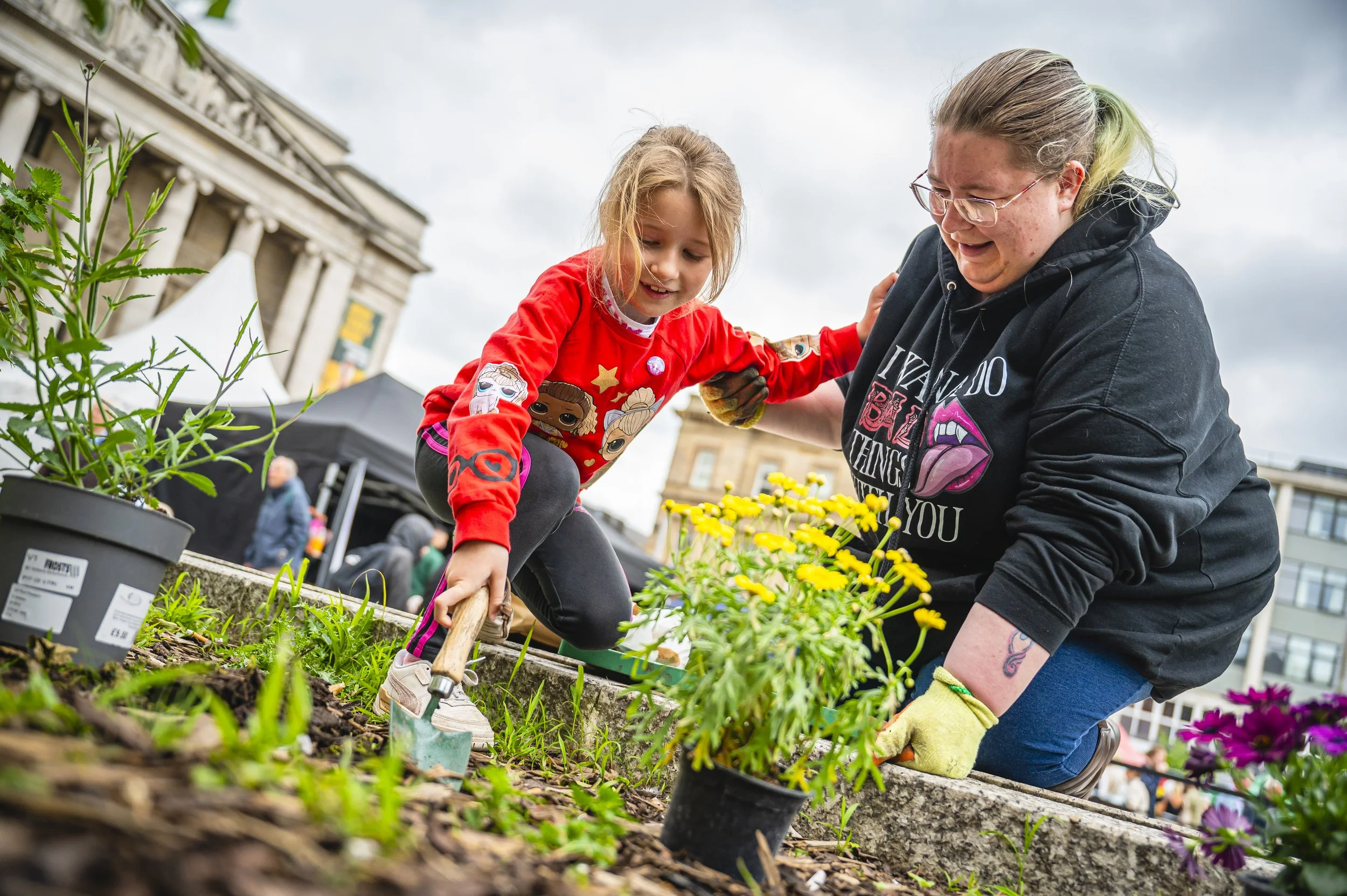 A young girl and woman gardening together outdoors, planting flowers in a public space with tents and buildings in the background.