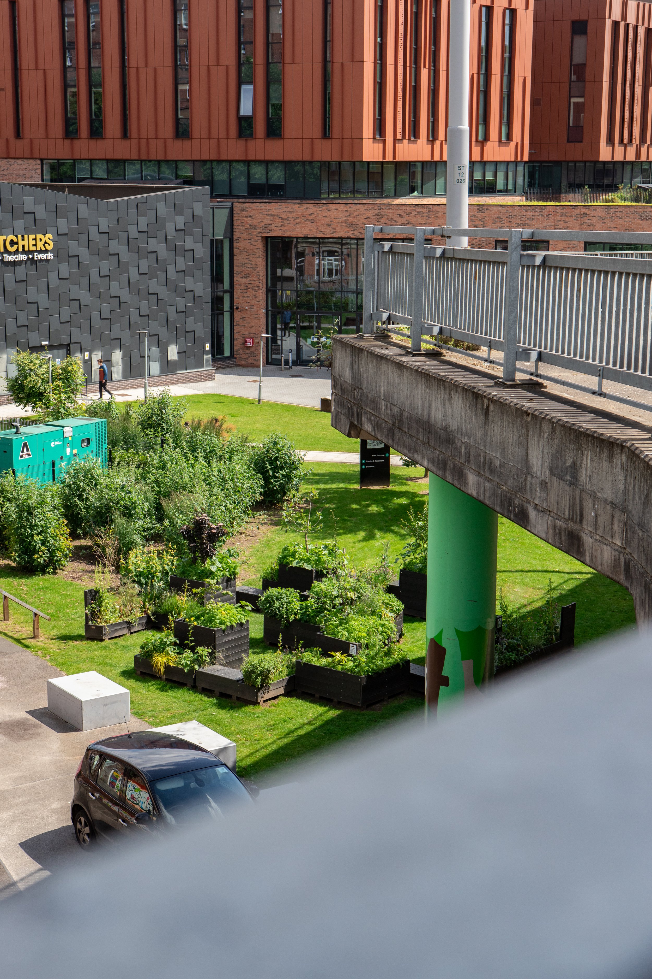 A cityscape view showing a landscaped area with planters filled with greenery, a parking area with a parked car, and a modern building with a textured dark gray exterior and large windows in the background. There is a bridge or elevated walkway with a railing and a green support pillar.